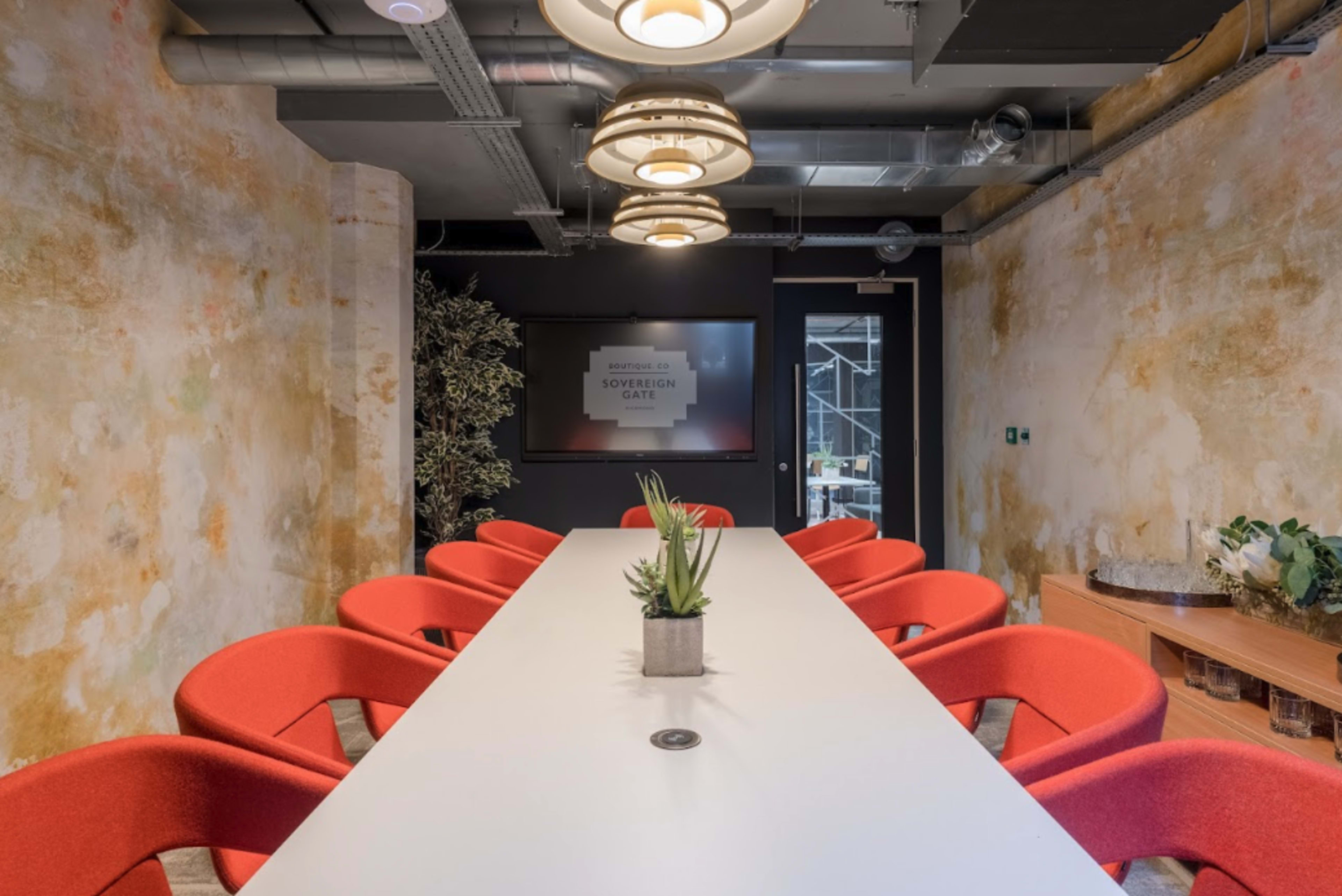 A modern conference room features a long white table surrounded by red chairs and an accent wall with a textured finish.