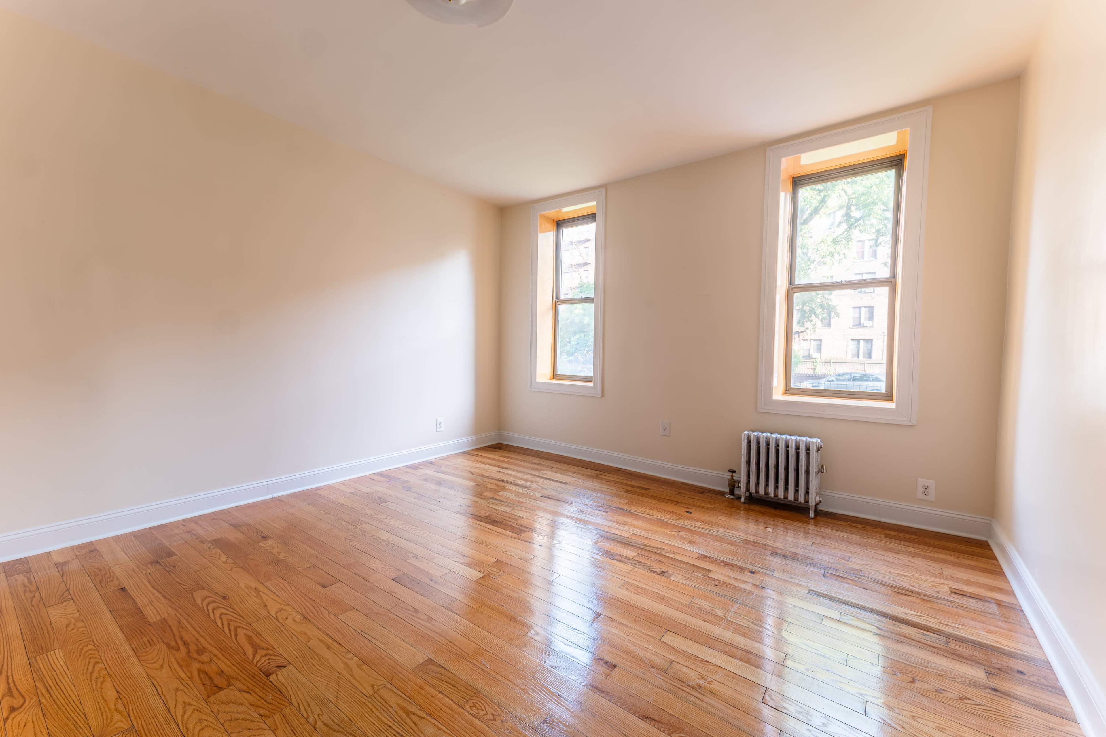 The image shows an empty room with two windows, hardwood floors, and a radiator.