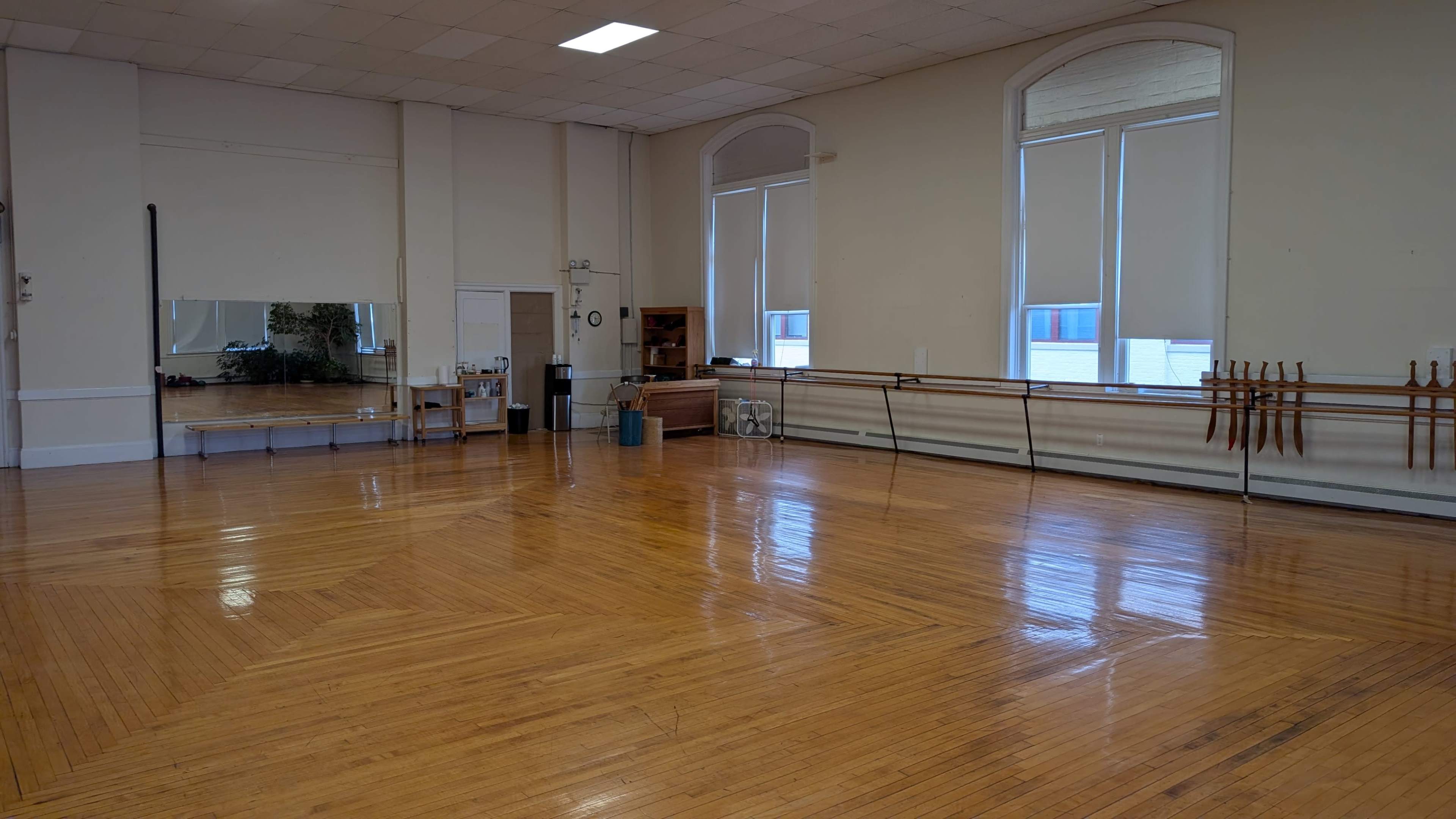 The image shows a spacious dance studio with polished wooden floors, large windows, and a mirror along one wall.