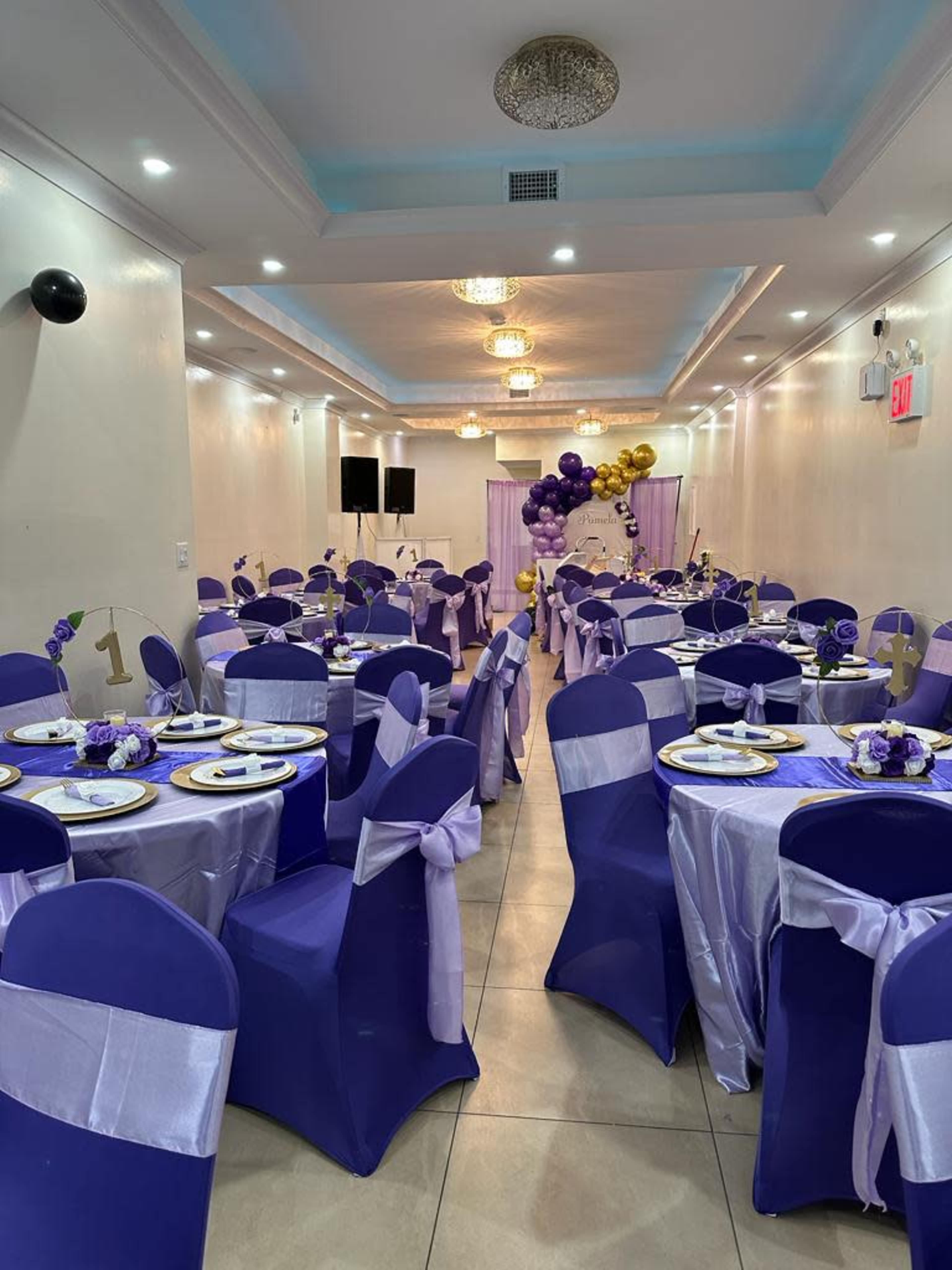 A decorated event space with tables set up in purple and white, featuring balloons and a stage area in the background.