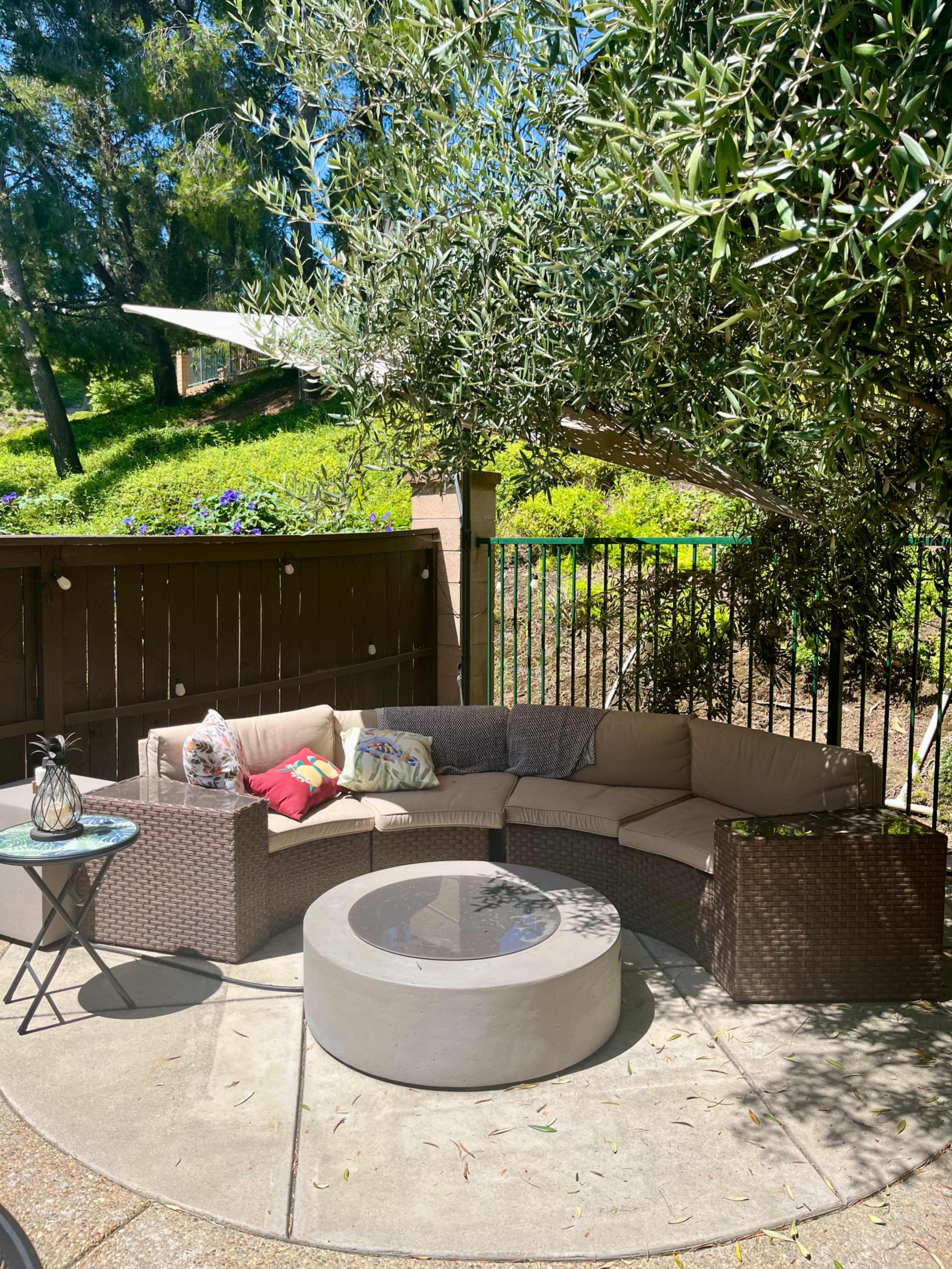 A circular outdoor seating area with a curved sectional sofa, a round coffee table, and a small side table is situated under a tree in a sunny garden.