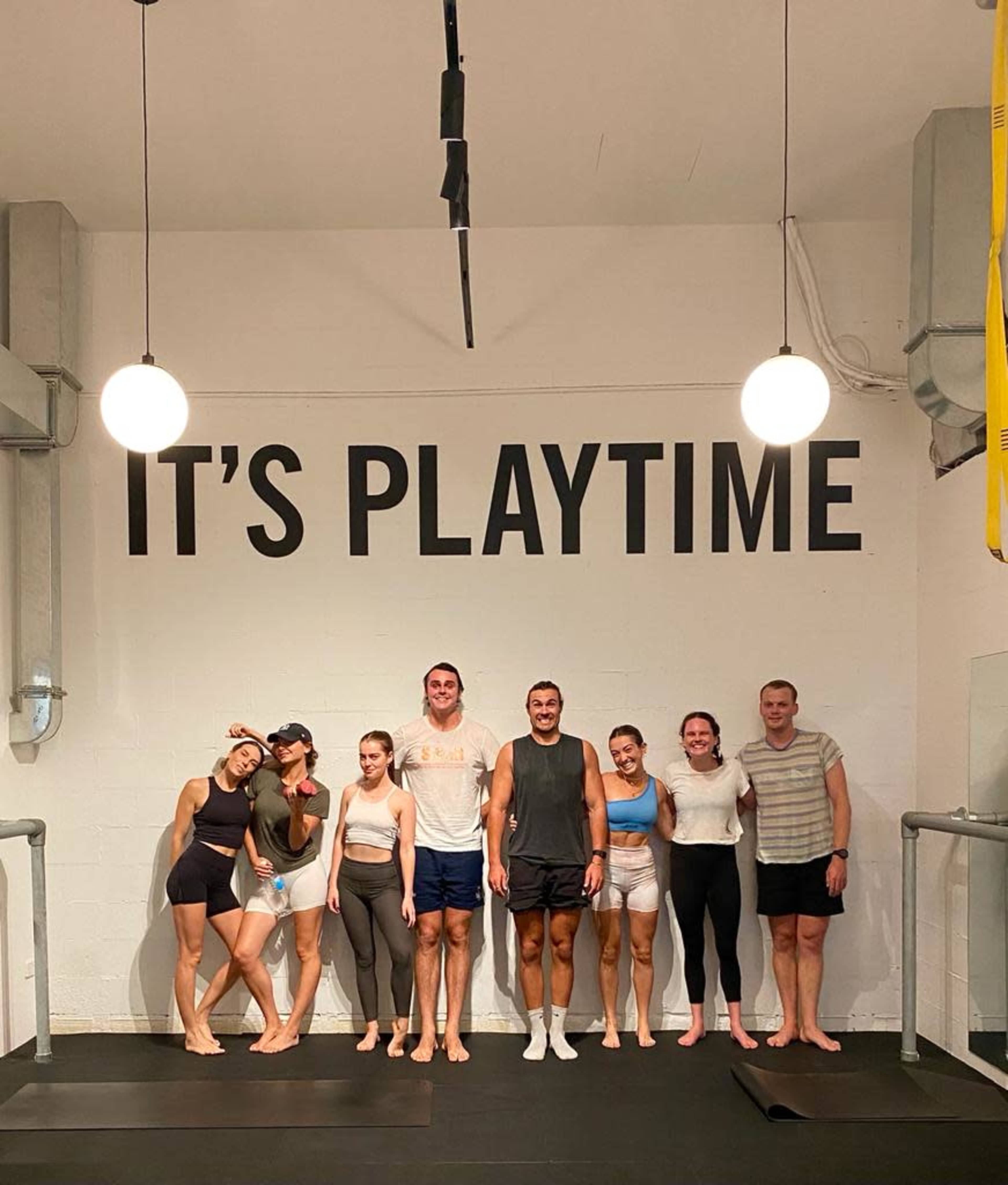A group of eight individuals poses together in a spacious room with a sign that reads "IT'S PLAYTIME" on the wall behind them.