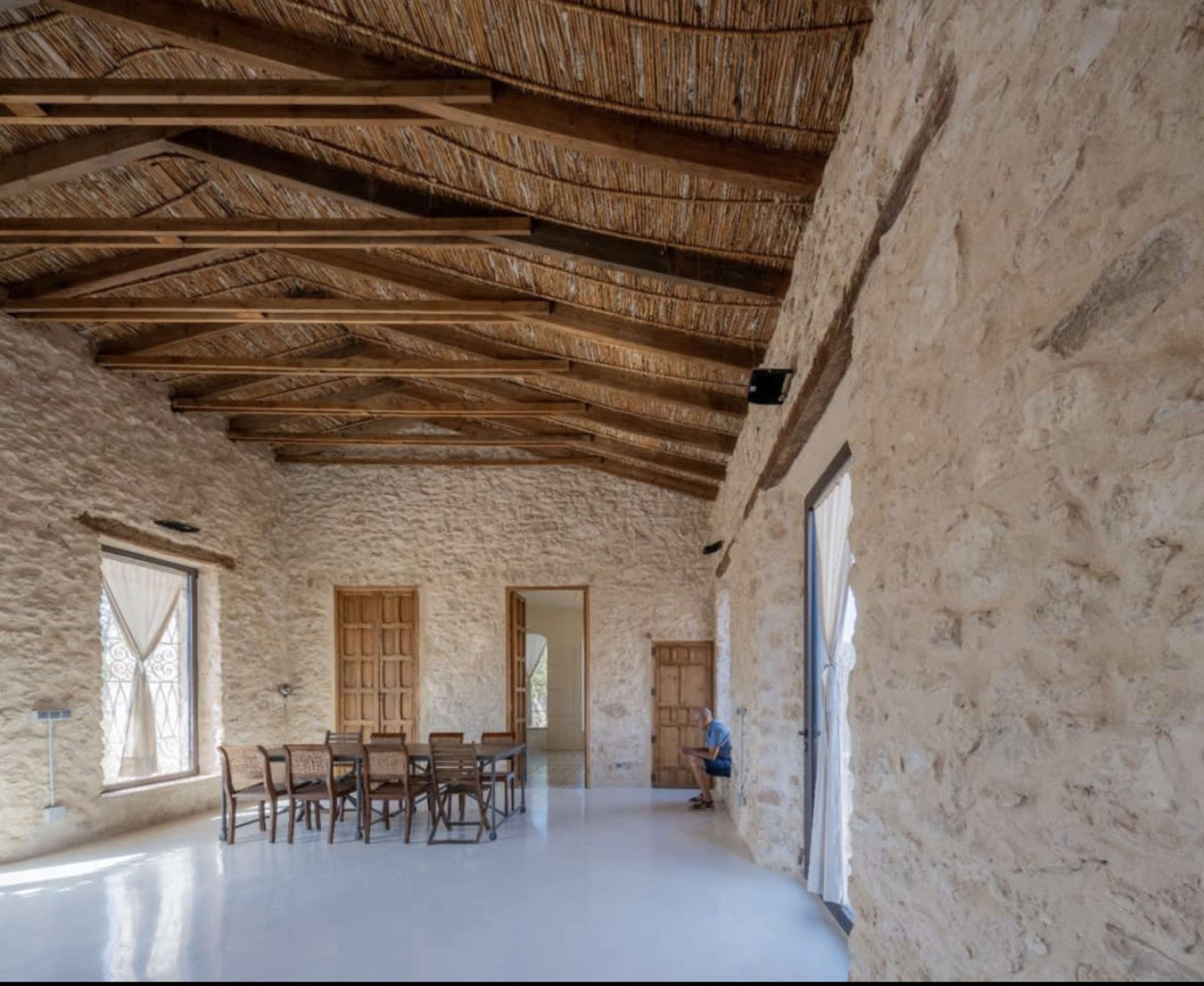 The image shows a spacious room with stone walls, wooden beams, a long dining table surrounded by chairs, and two large windows covered with white curtains.