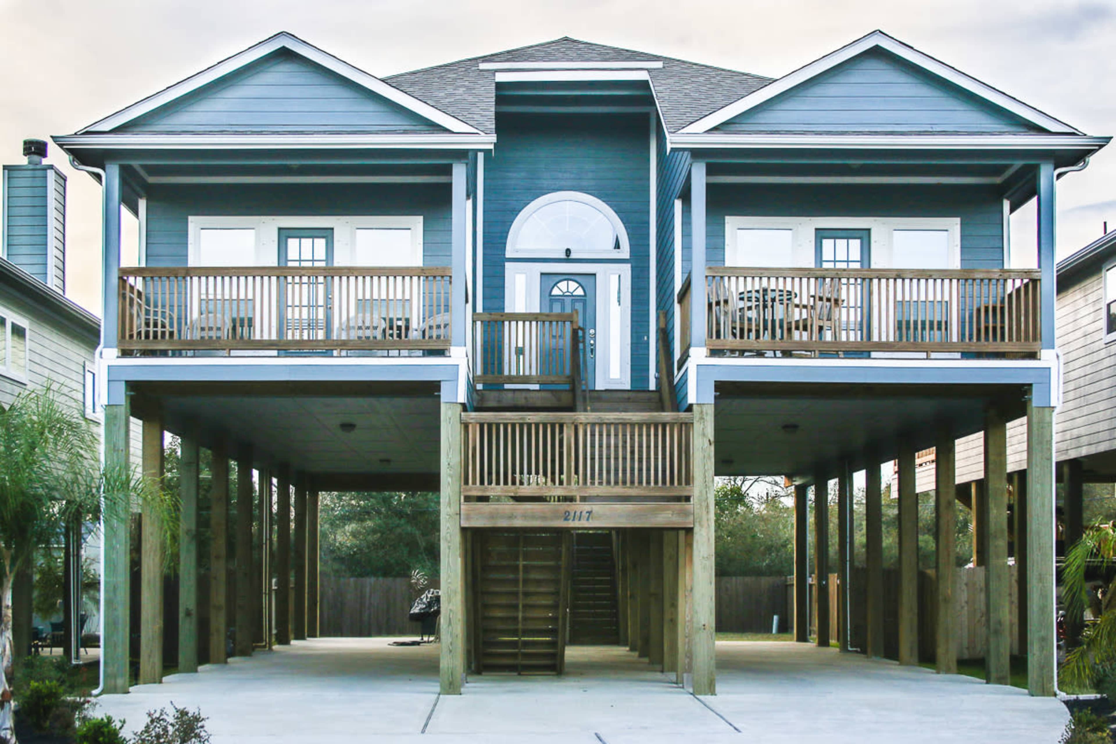 A two-story blue house with a central front entrance and balconies on both sides is supported by wooden stilts.