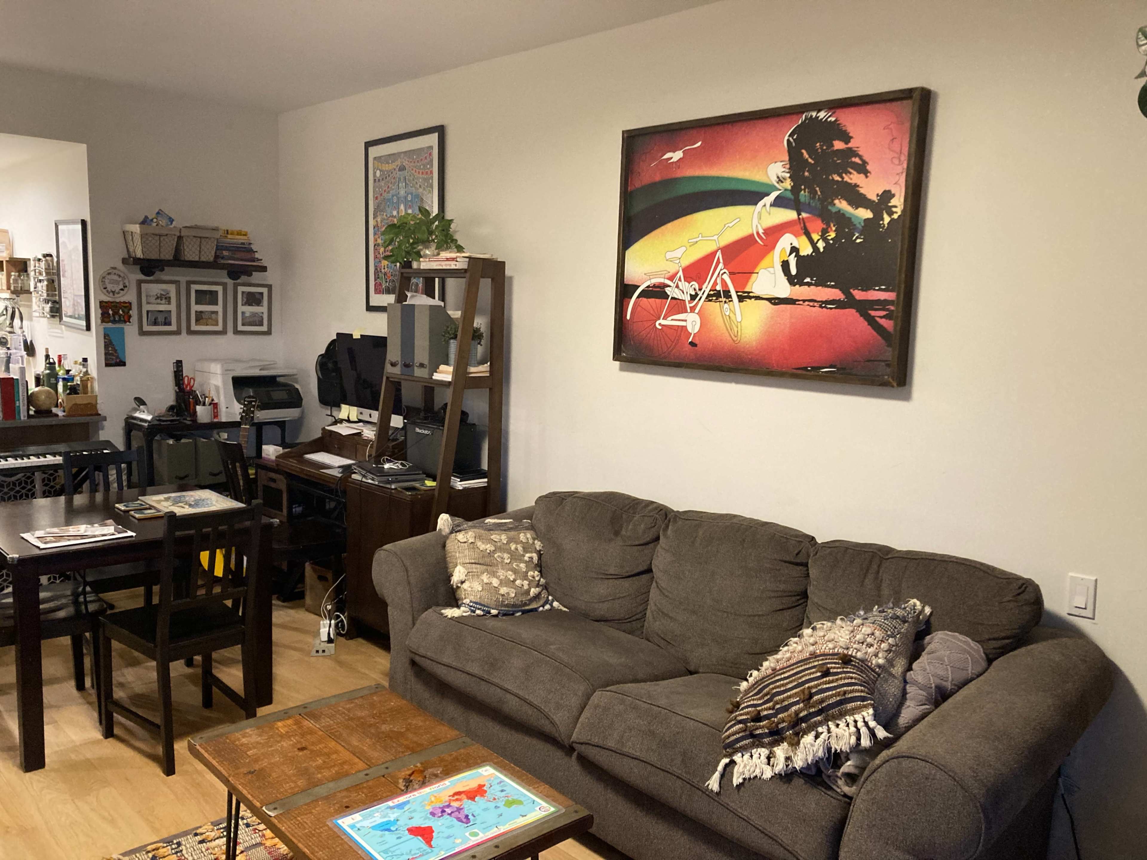 The image shows a living room with a gray couch, a wooden coffee table, a dining area with a black table and chairs, and artwork featuring a beach scene on the wall.