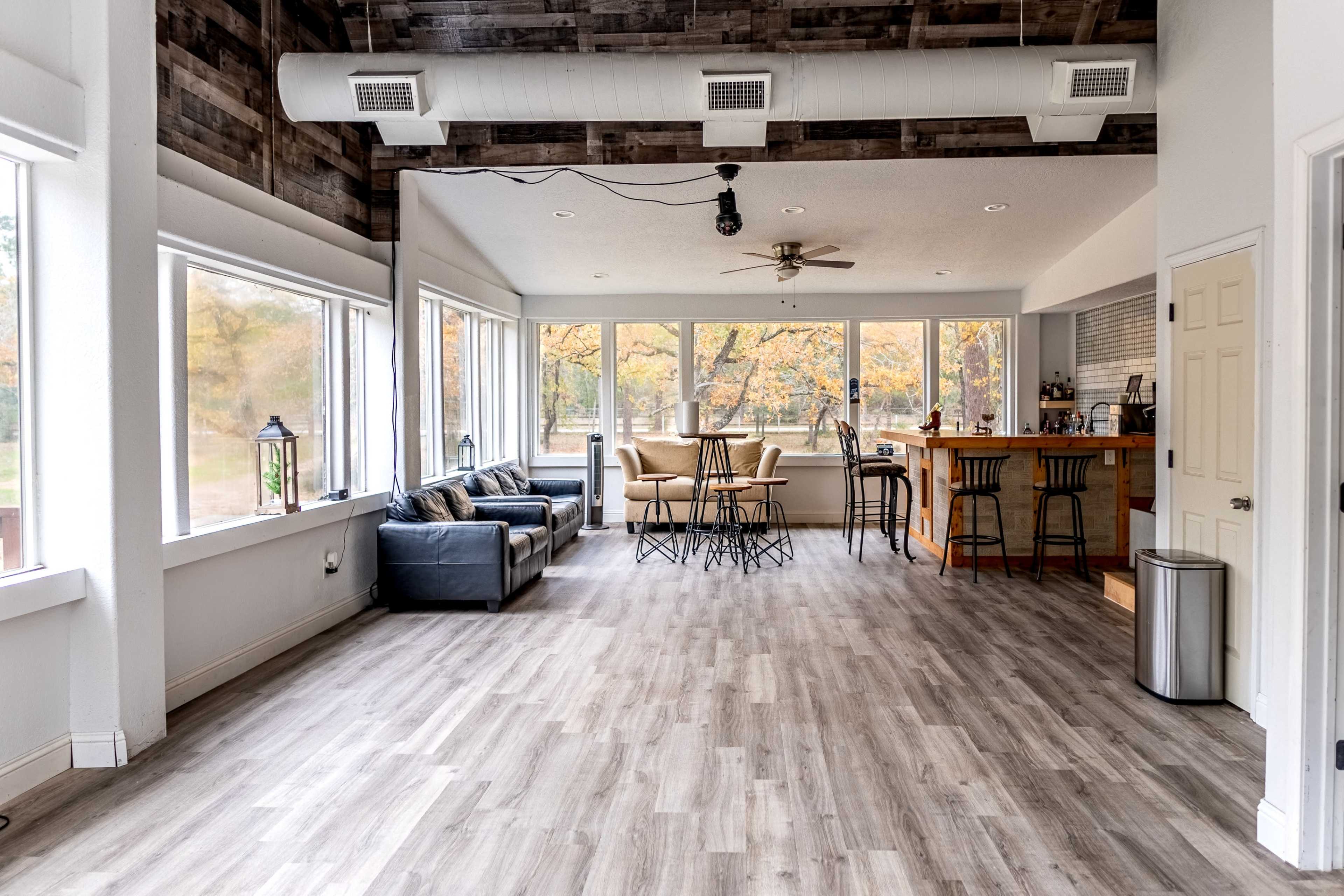 The image shows a spacious, modern living area with large windows, a seating area, a dining table, and a kitchen counter.