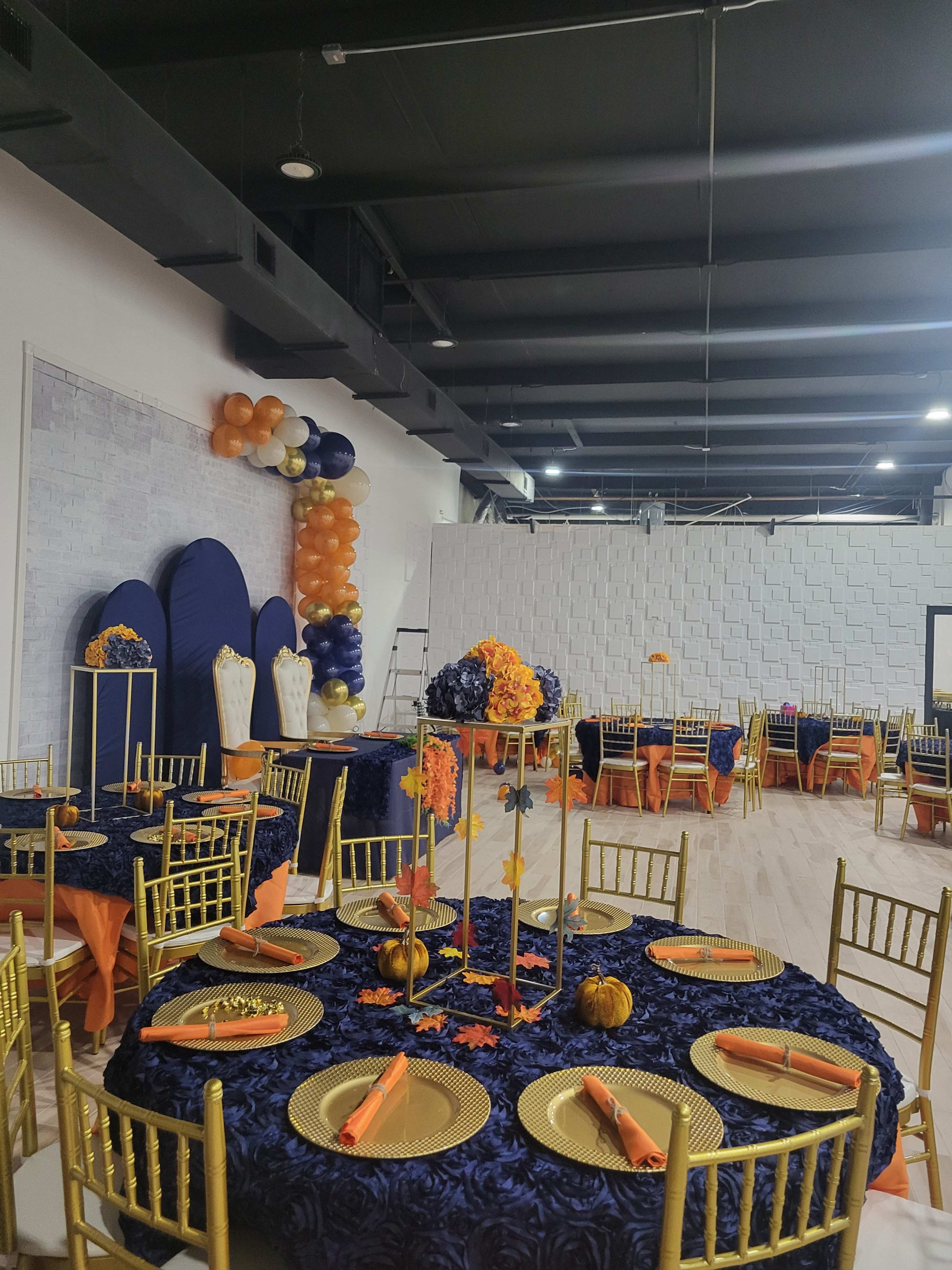 The image shows a banquet hall decorated for an event, featuring tables covered with navy blue tablecloths, gold chairs, and orange accents, with a backdrop of balloons and floral arrangements.
