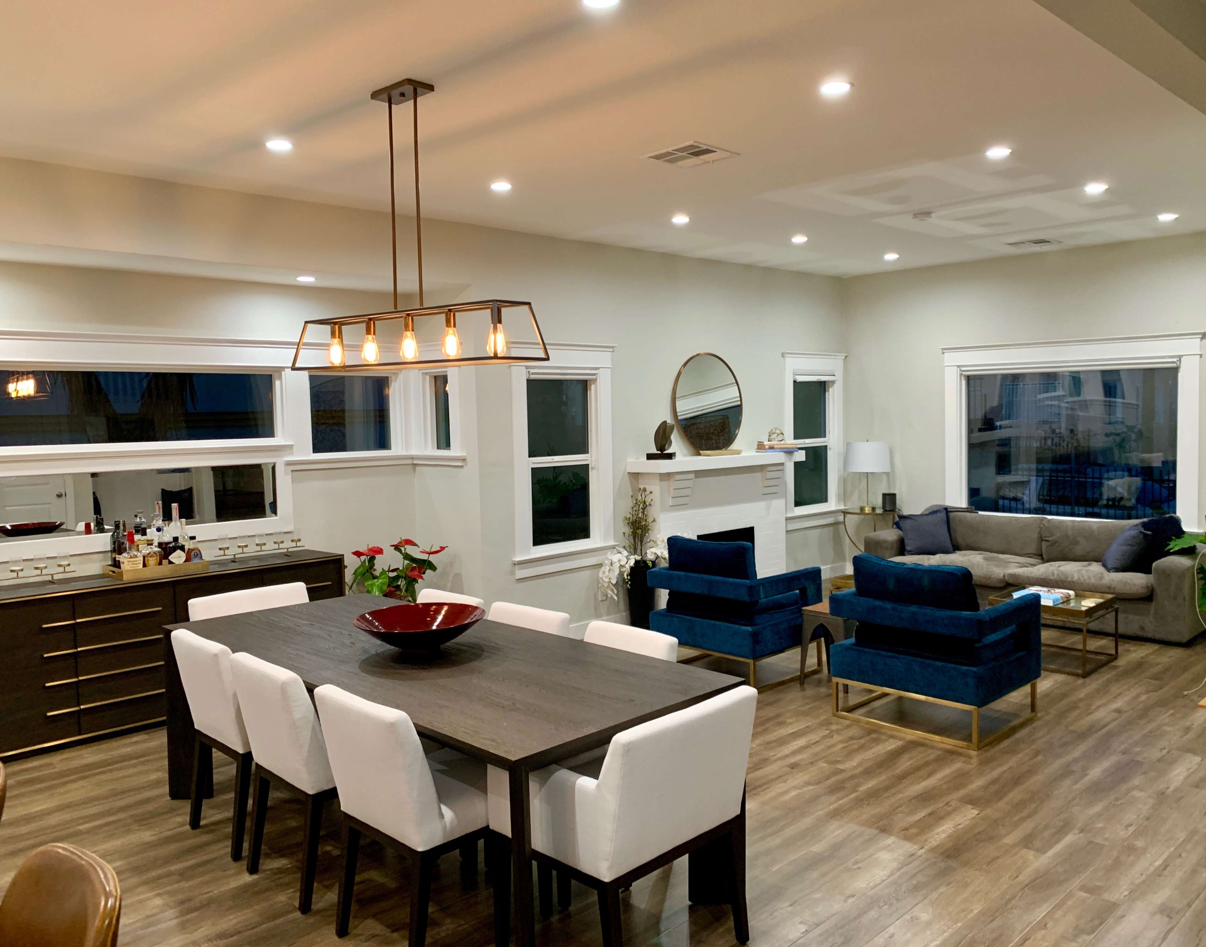 The image shows a modern living and dining area featuring a long dining table with white chairs, a dark sideboard, and a cozy seating arrangement with blue sofas and a round mirror above a fireplace.