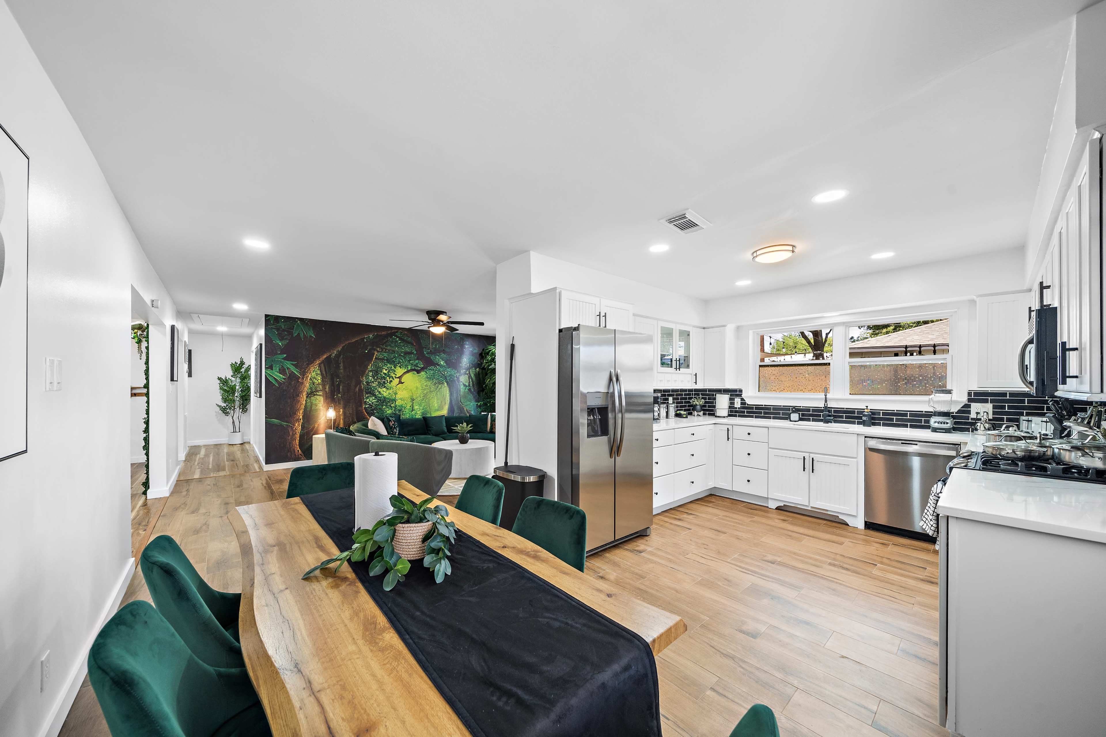 A modern kitchen and dining area featuring a wooden dining table with dark green chairs, stainless steel appliances, and a mural of a forest on one wall.