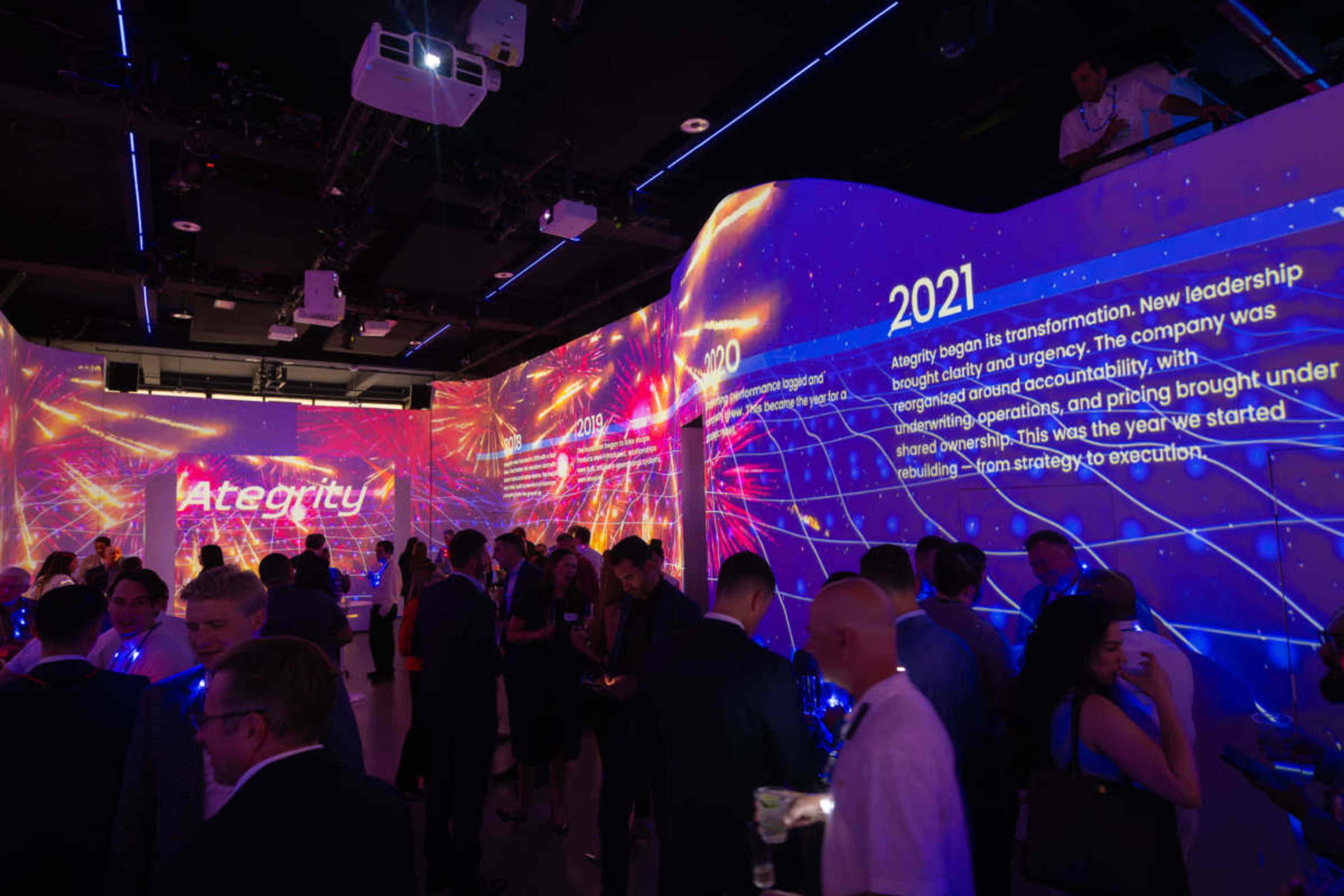 The image shows a lively event space featuring large, illuminated screens displaying information and graphics, with attendees mingling and engaging in conversation.