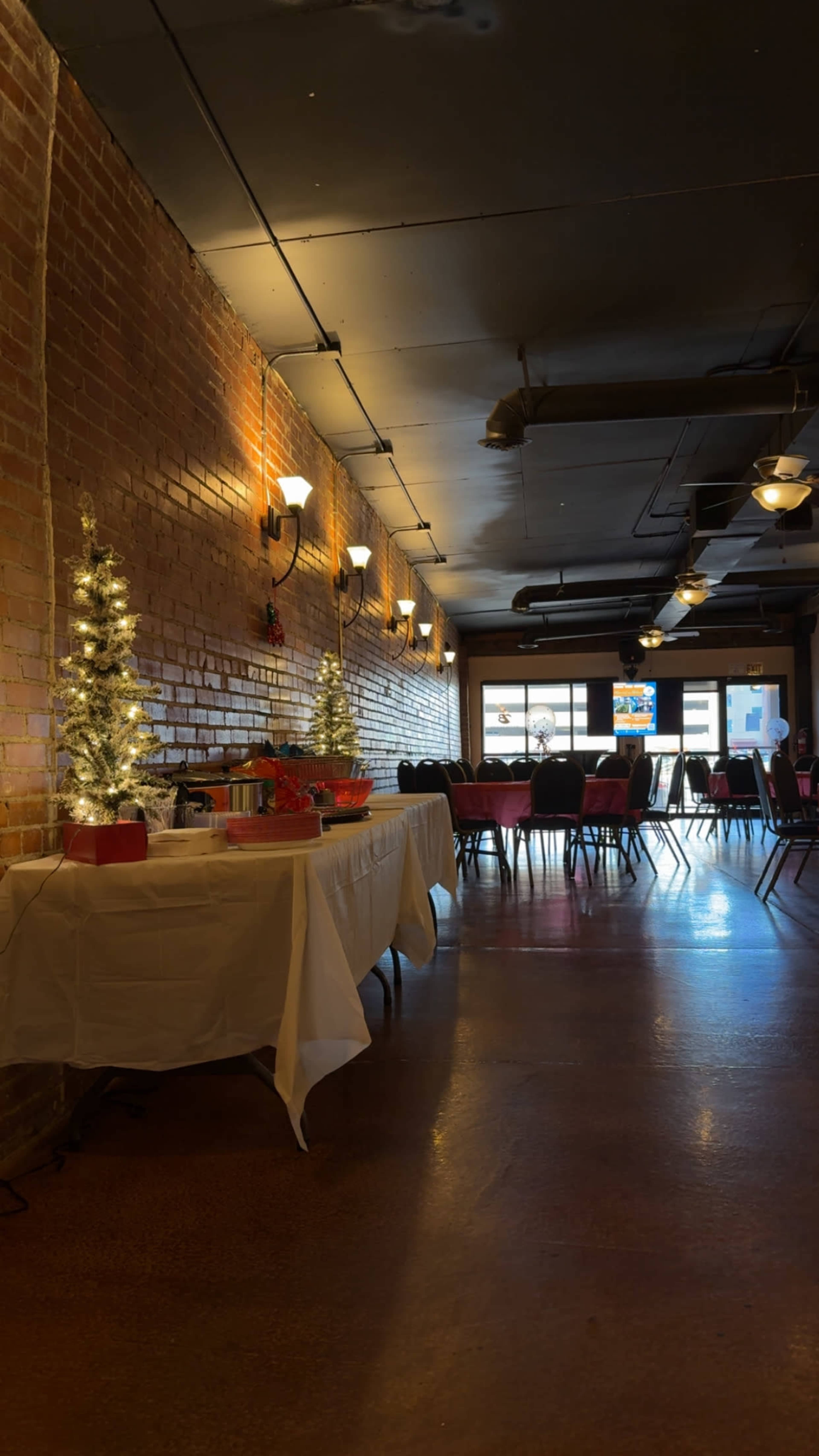 The image shows a festive indoor space with small Christmas trees and a table set up, alongside rows of chairs and tables in preparation for an event.