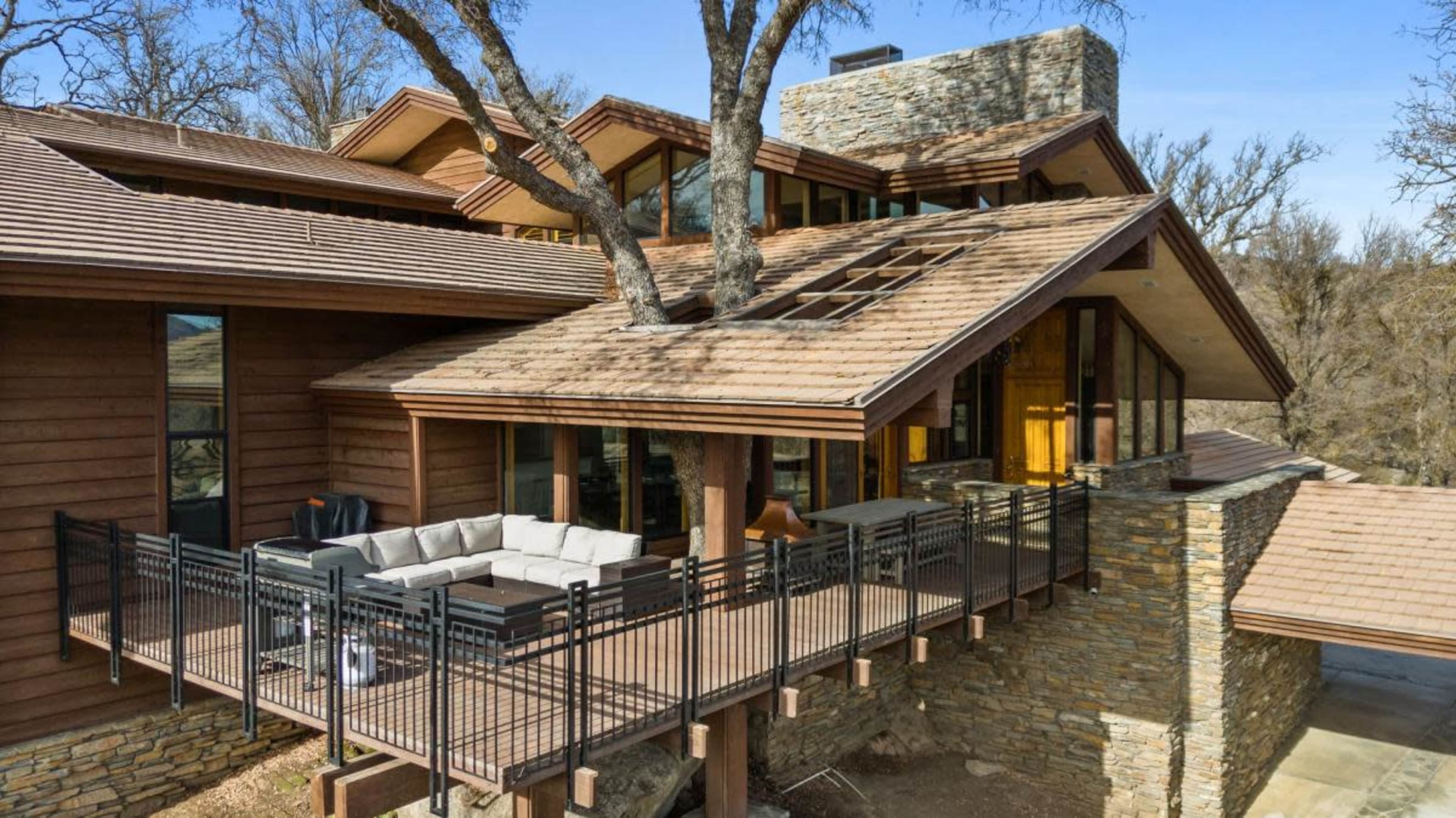 A modern house with a combination of wood and stone exteriors, featuring a large deck with outdoor seating and a tree growing through the roof.