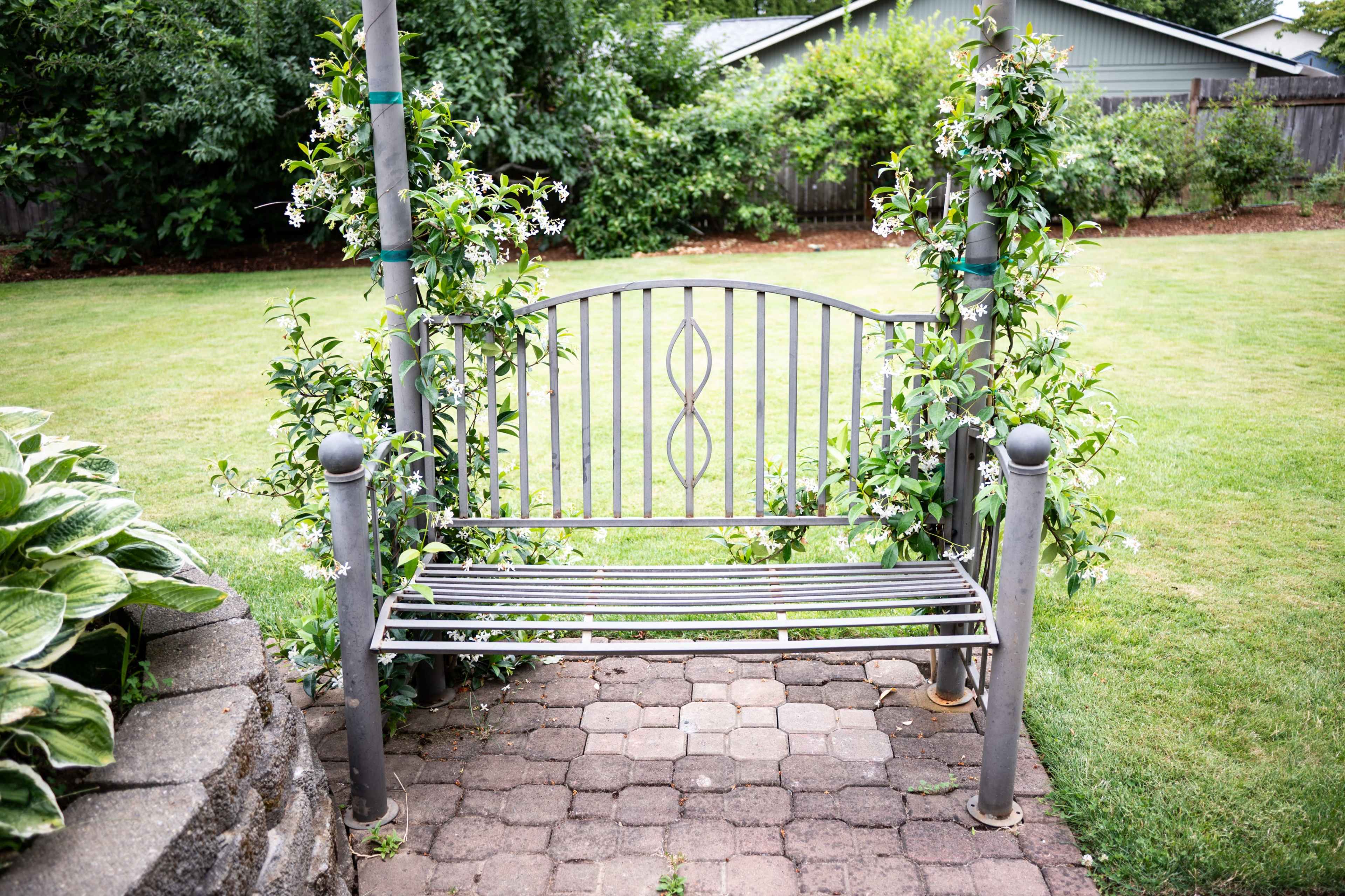 A metal bench surrounded by lush green plants is set in a garden area with a stone pathway.