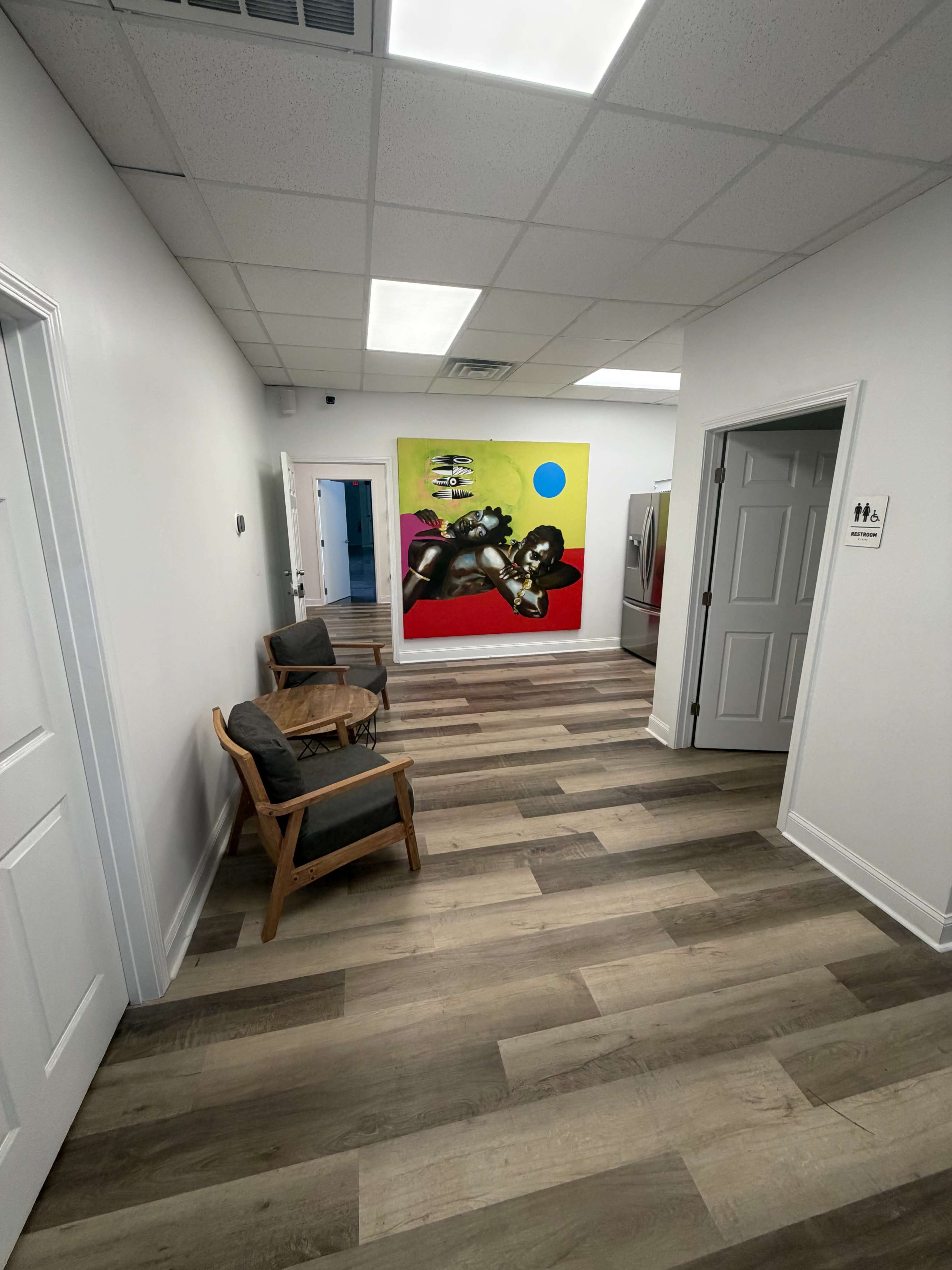The image shows a hallway with light-colored walls, wooden flooring, and a colorful mural on one wall, featuring a dynamic figure, along with two doors on the right and a seating area with two wooden chairs on the left.
