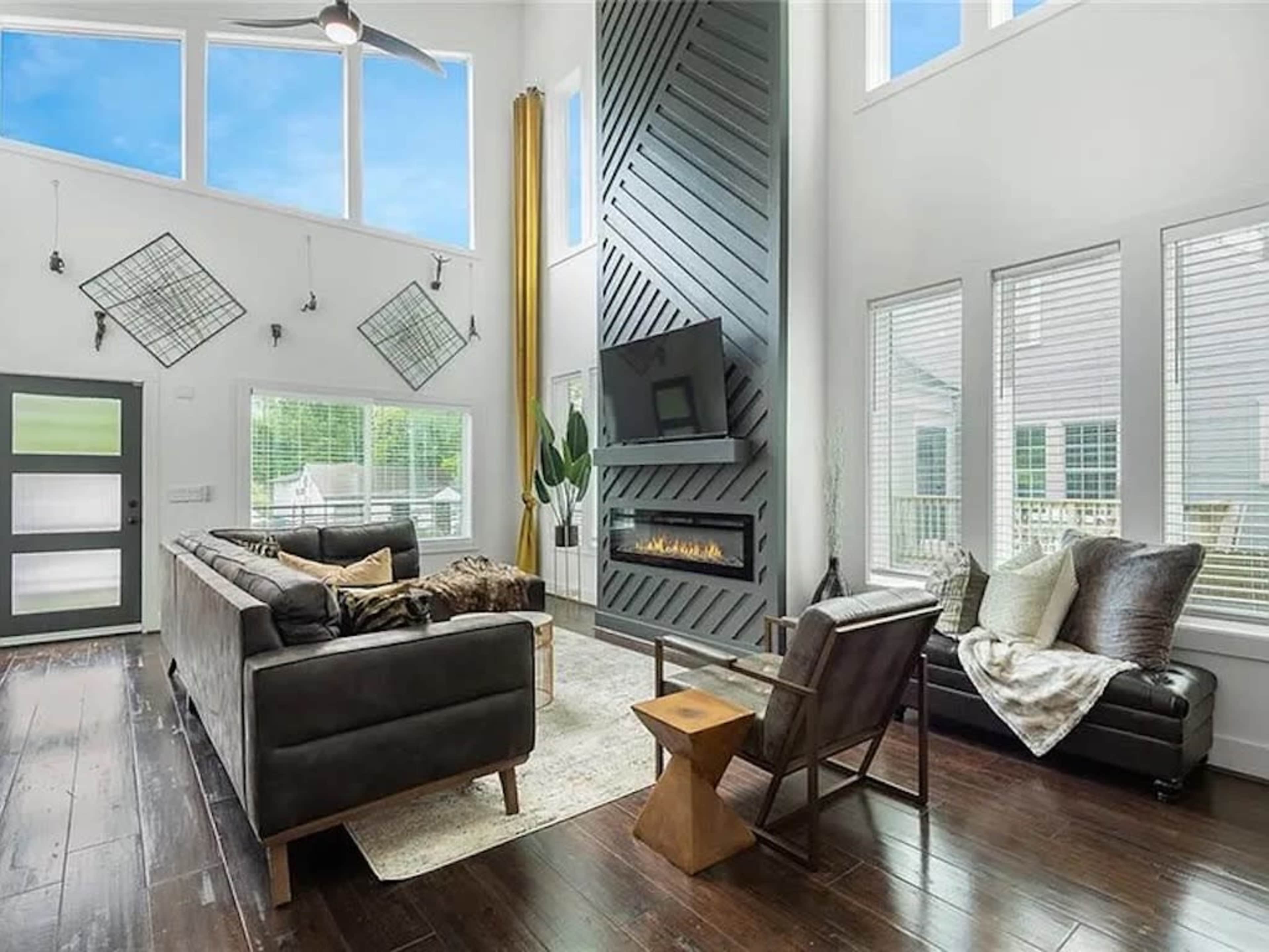 A spacious living room features a tall ceiling, large windows, a modern fireplace, and sectional seating arranged around a central rug.