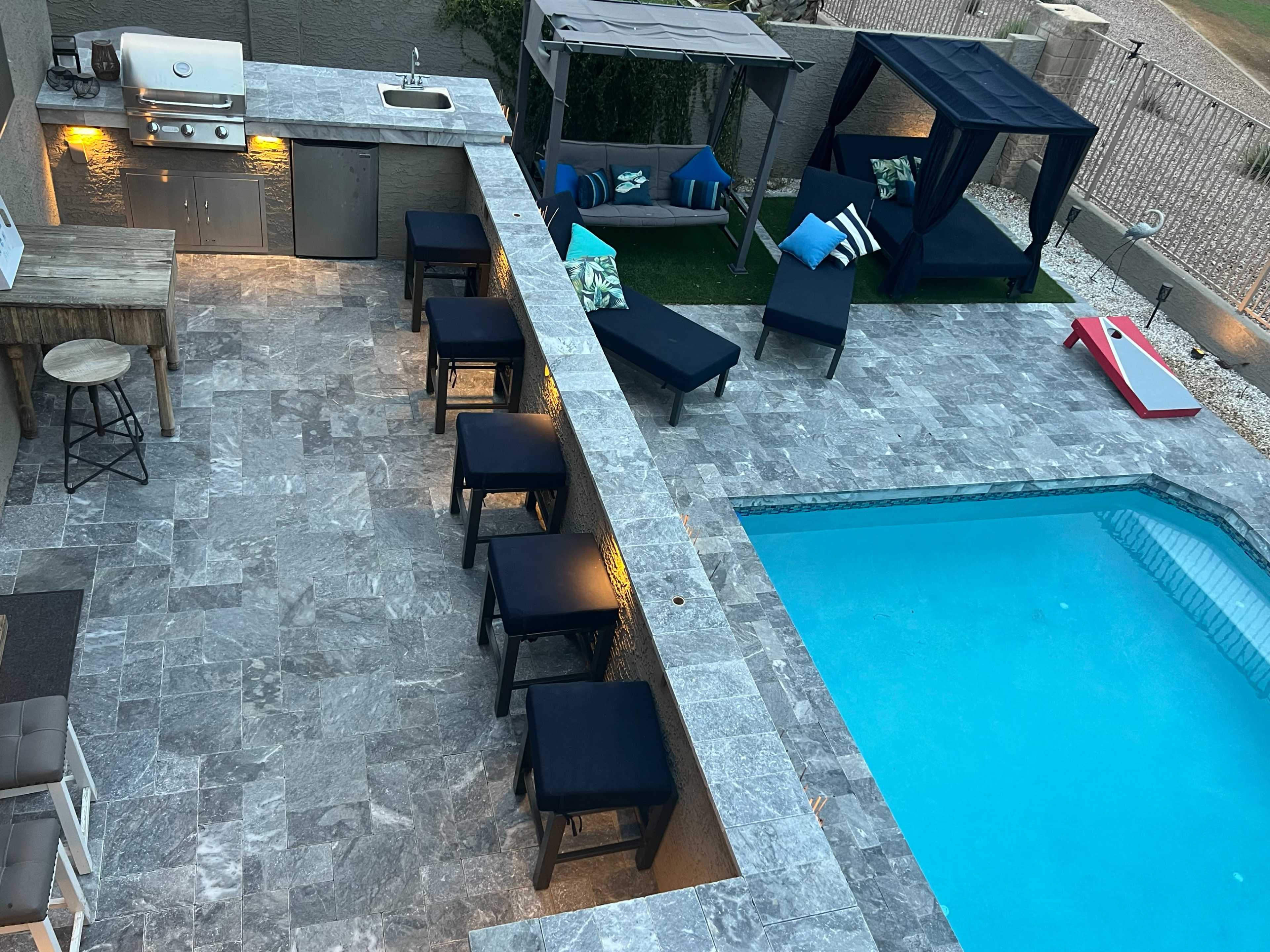 The image shows an outdoor patio area featuring a swimming pool, a bar with high stools, a grill, and two shaded seating areas.