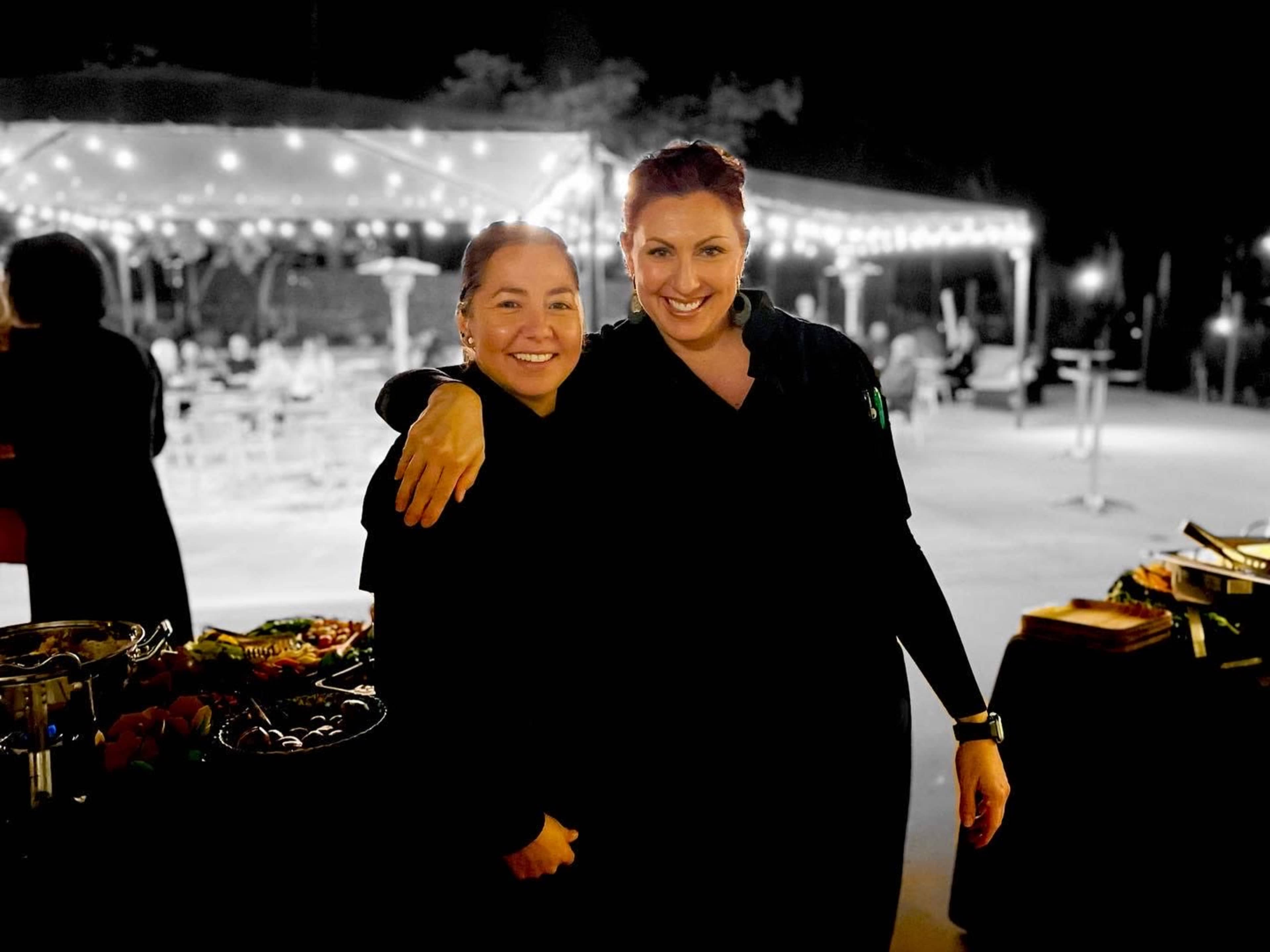 Two women in black attire stand closely together, smiling in front of a festively lit outdoor event setting.