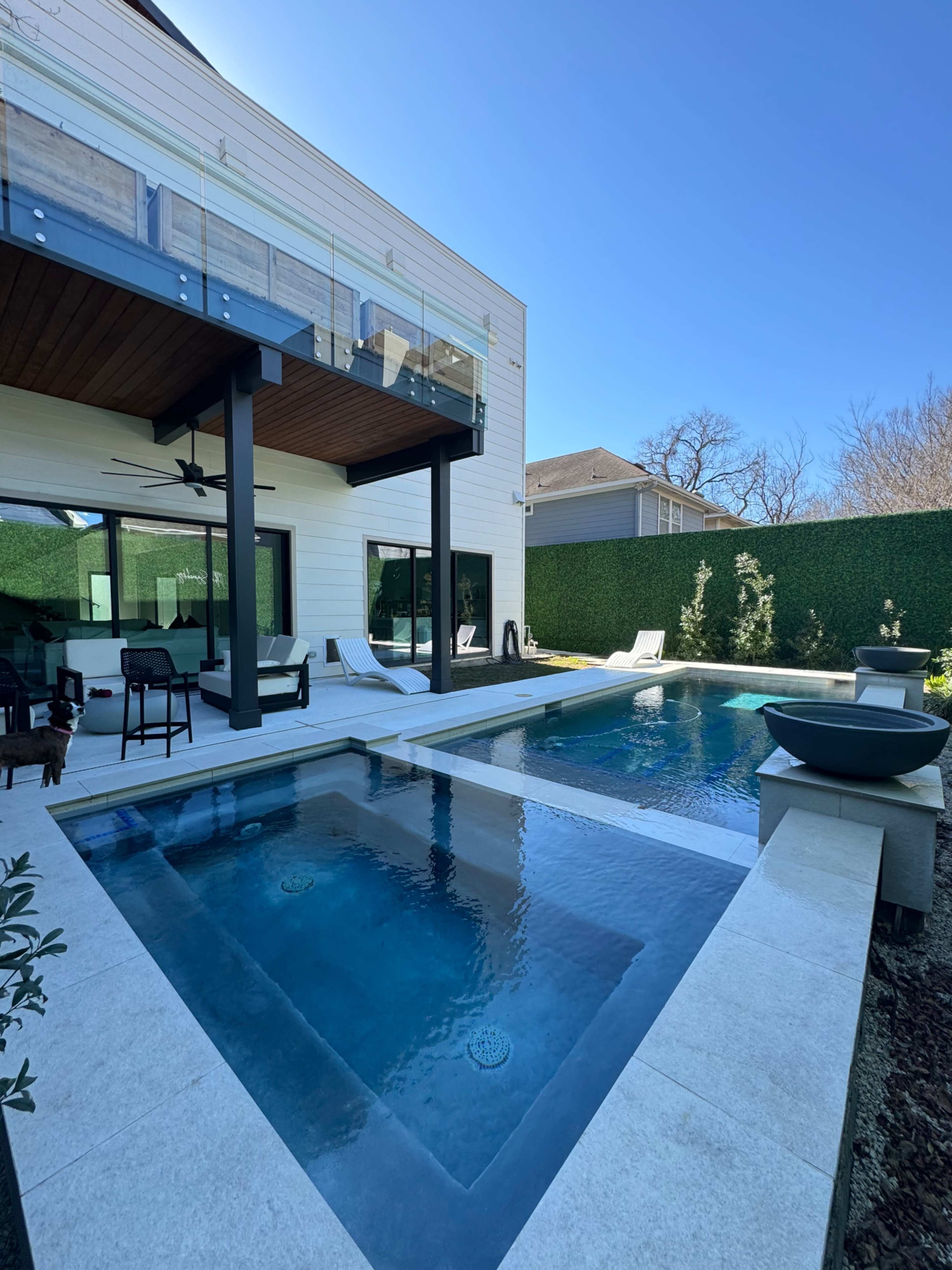 A modern outdoor area featuring a pool, a patio with seating, and lush green hedges surrounding the space.