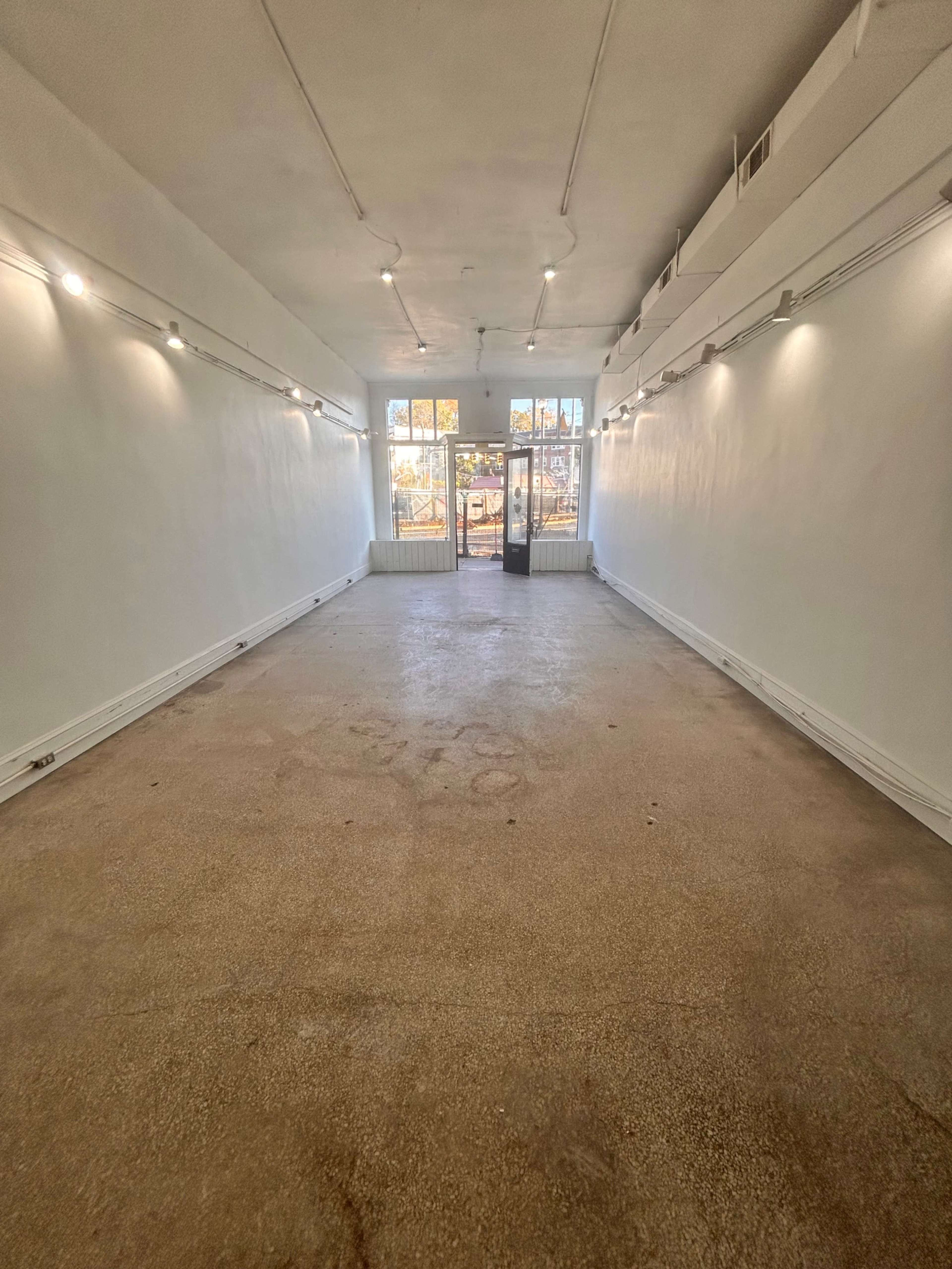 The image shows a spacious, empty room with white walls, bright lights, and large windows leading to an outdoor area.