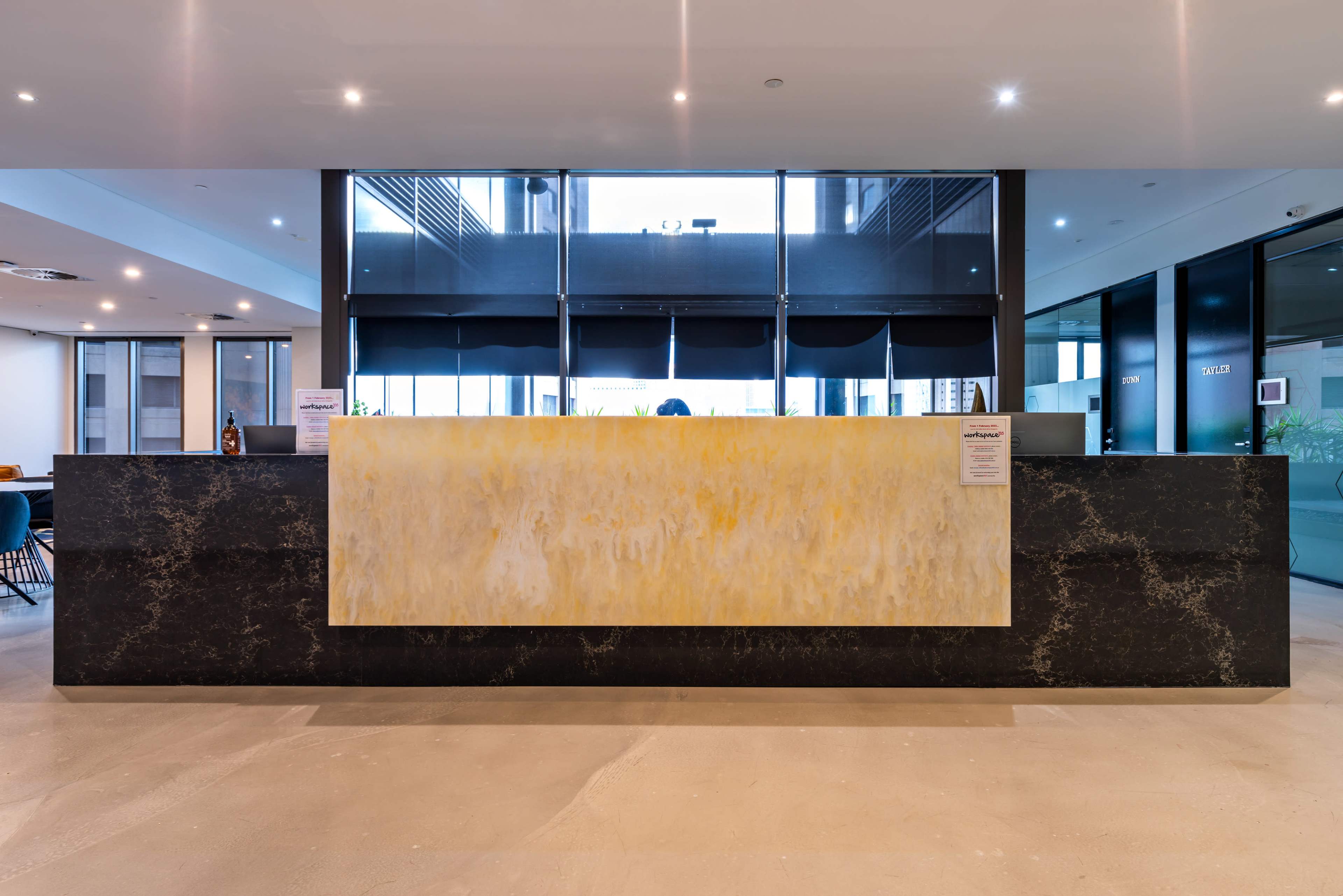 The image shows a modern reception desk made of two contrasting materials, with a light-colored panel on top of a darker base, situated in a well-lit lobby area featuring large windows.