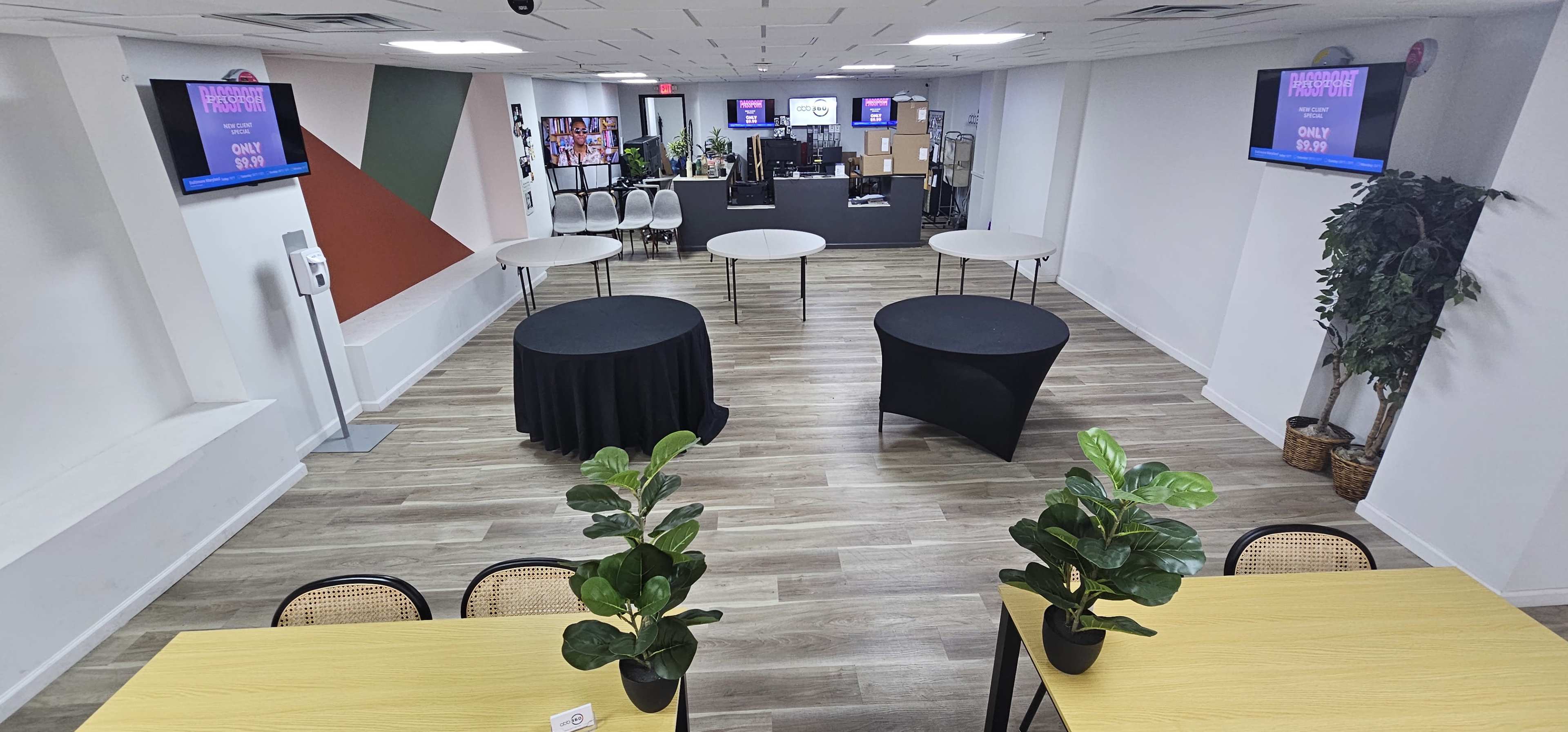 The image shows a spacious, modern event room featuring several tables, plants, and multiple screens displaying event information.