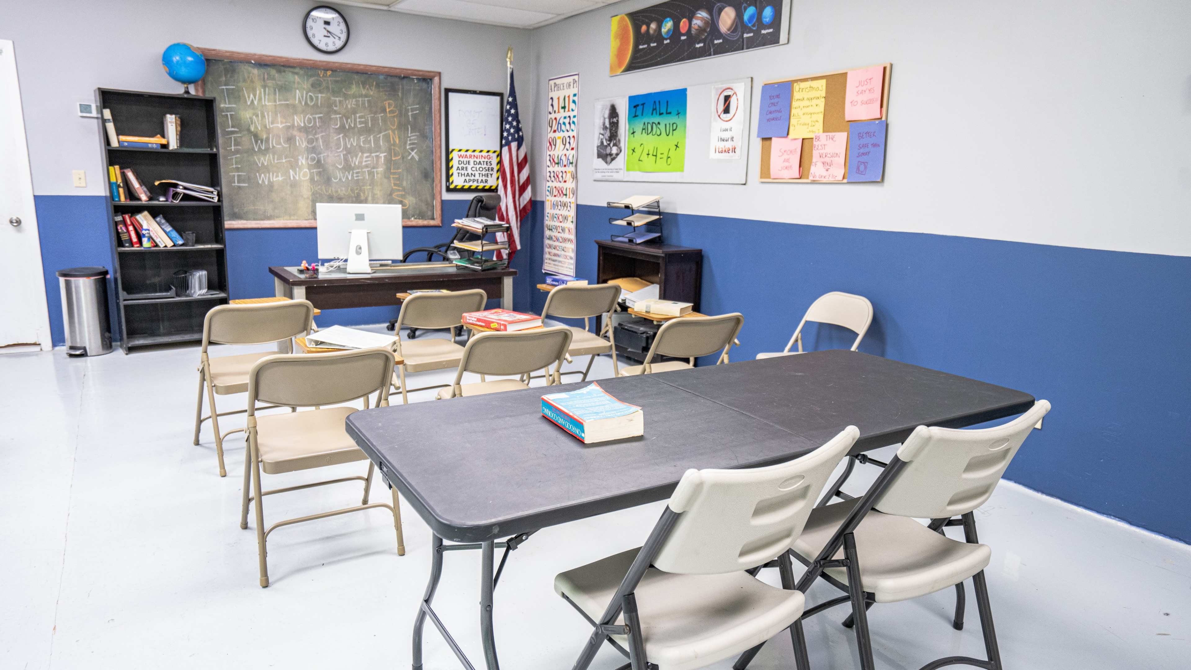 The image shows a classroom with a blackboard, desks arranged in rows, a computer, and educational posters on the walls.
