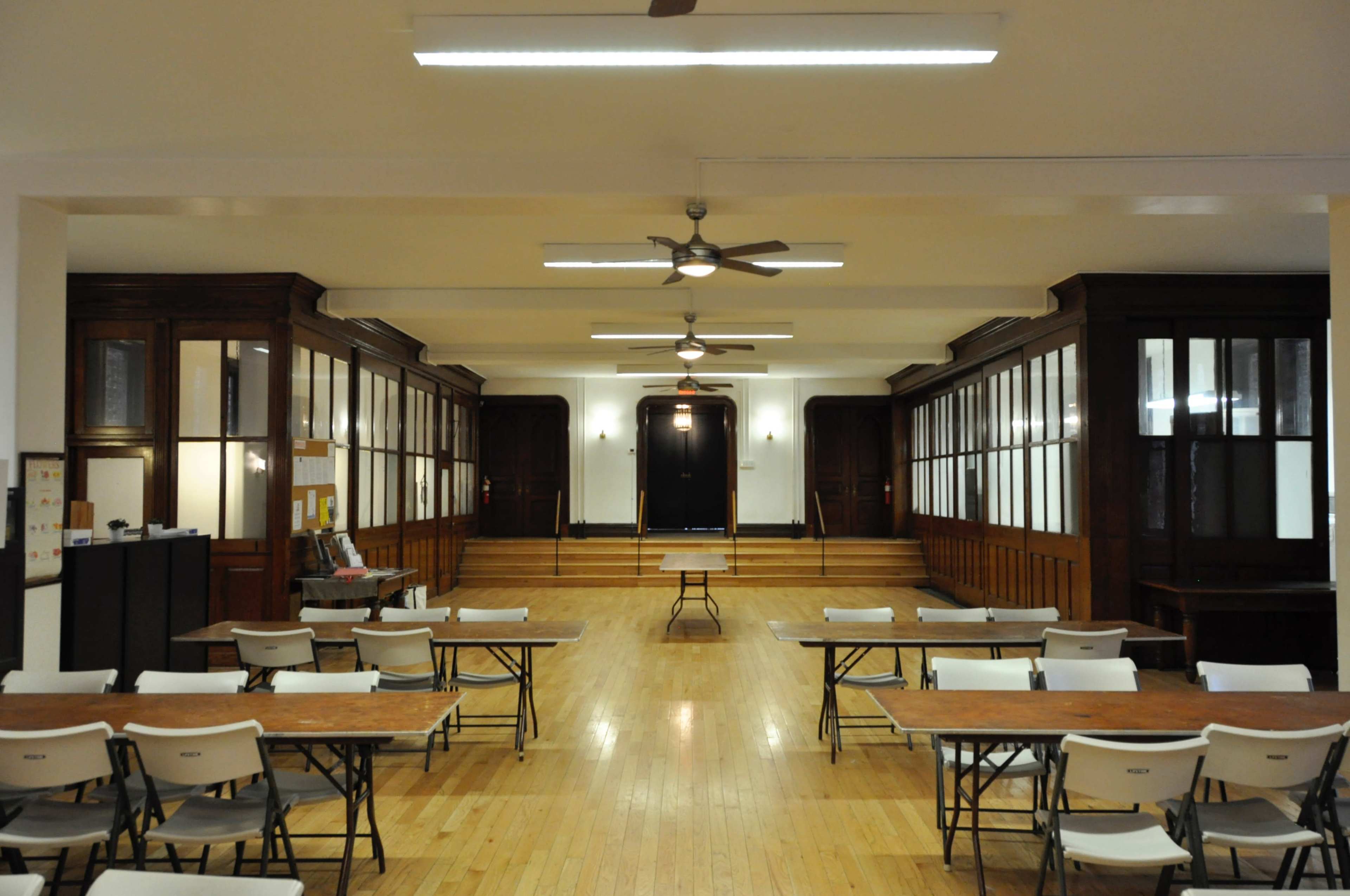 A large, empty room with several tables and chairs arranged in rows, flanked by wooden walls with glass panels.
