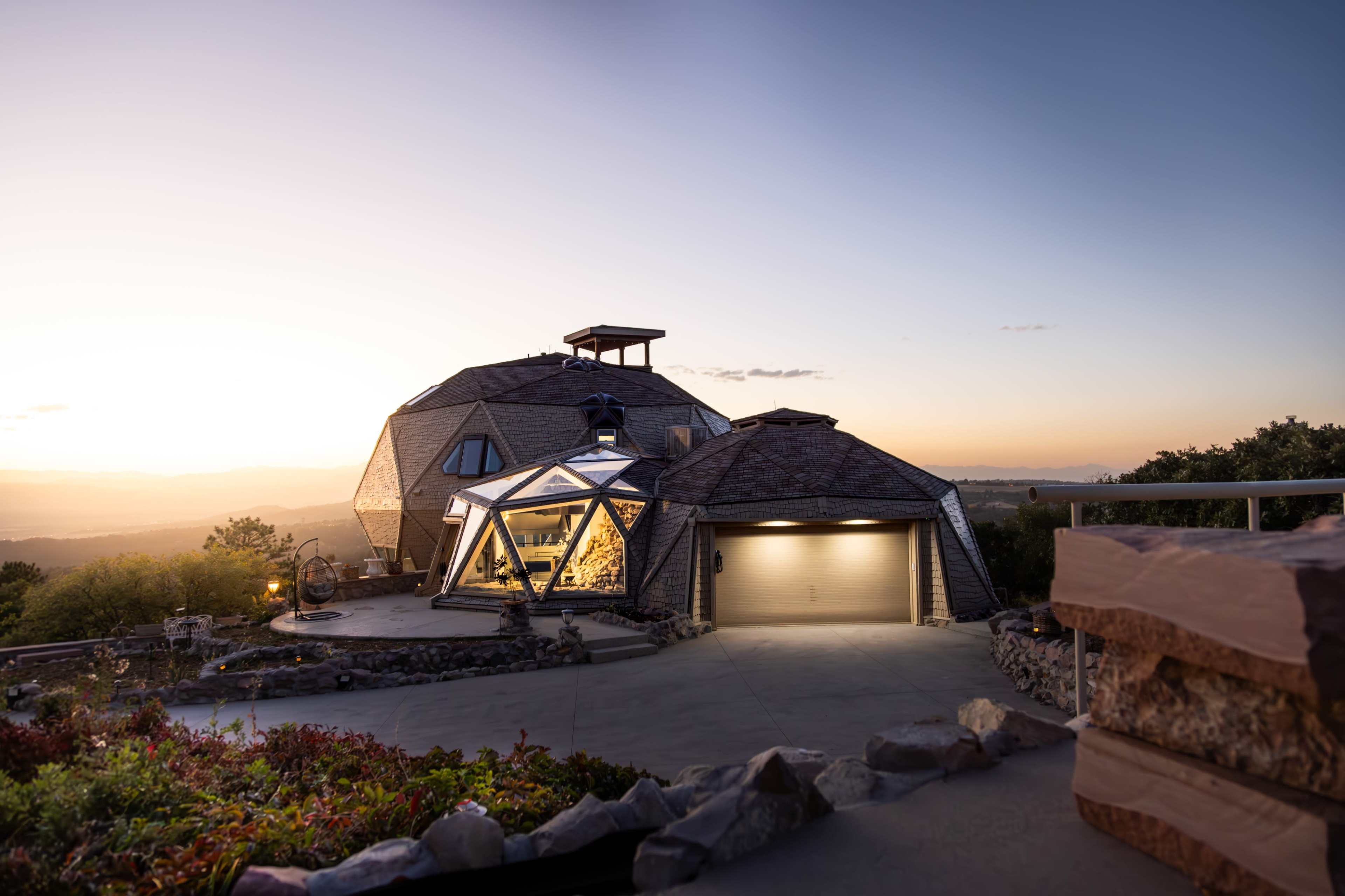 A modern geodesic dome house sits on a hillside at sunset, showcasing its glass and stone exterior.
