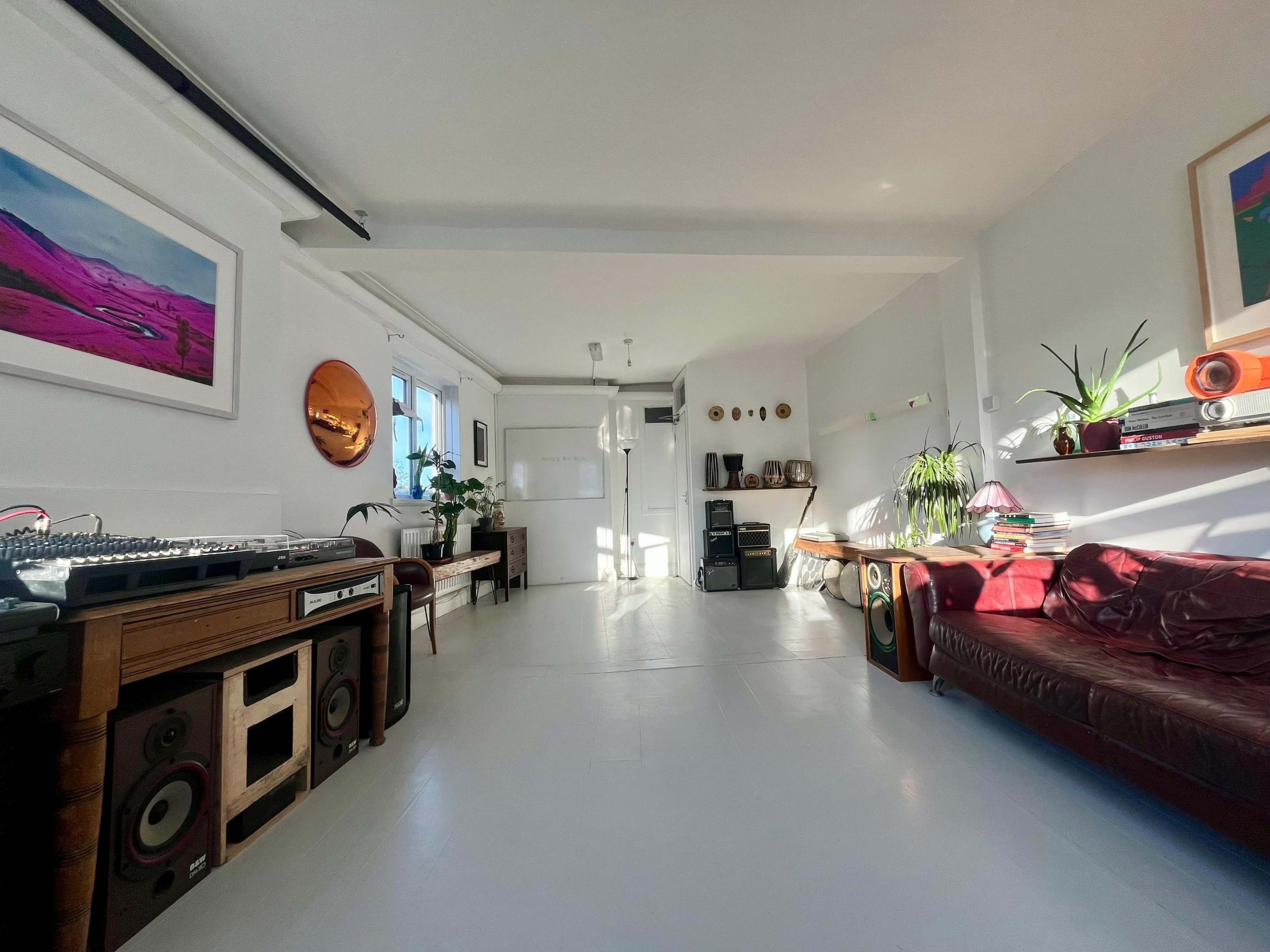 The image shows a spacious, well-lit room featuring a sofa, a table with a sound mixing console, and decorative plants along with colorful artwork on the walls.