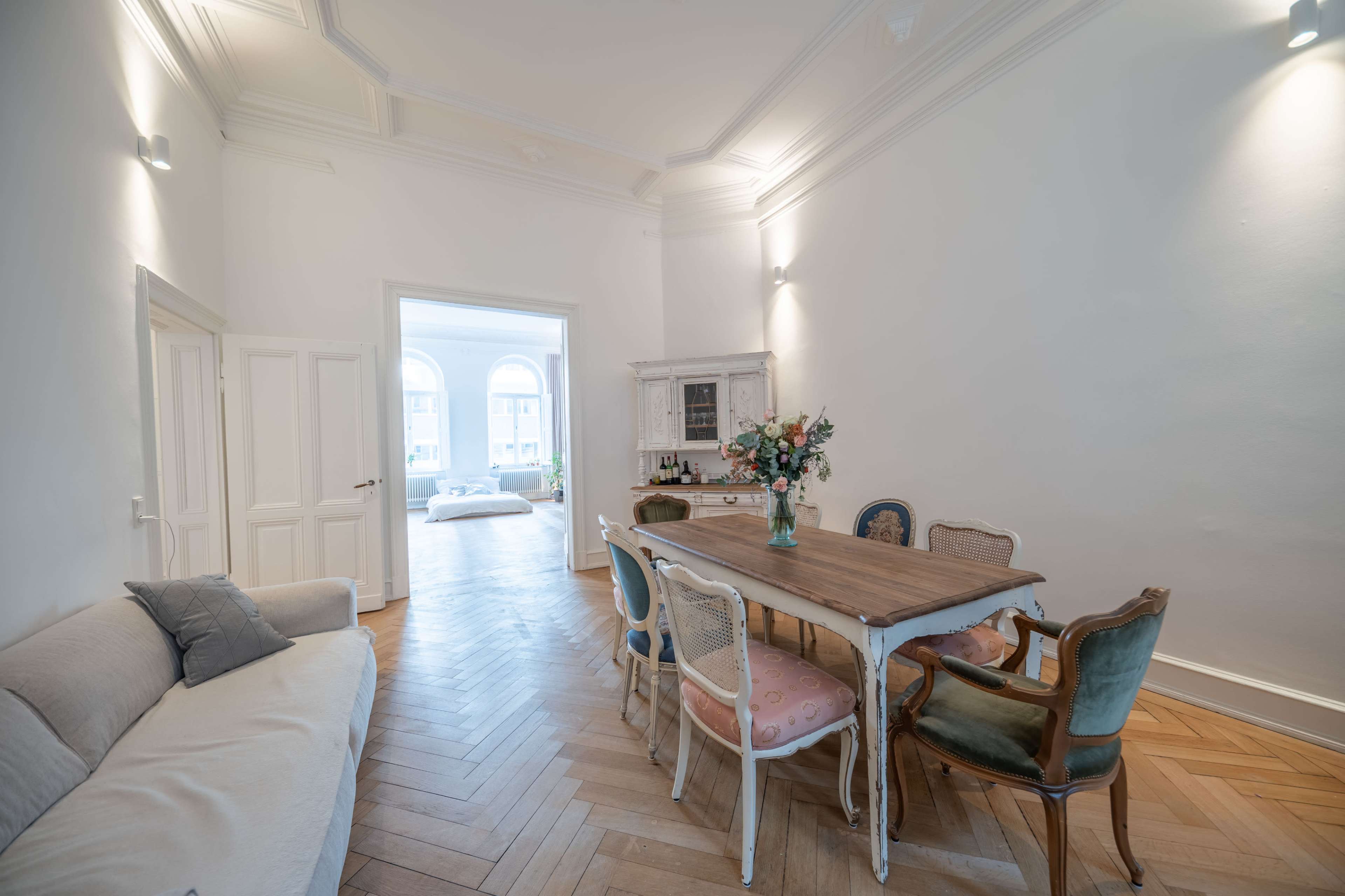 The image shows a bright room with a wooden dining table surrounded by chairs, a sofa against one wall, and a doorway leading to another area.