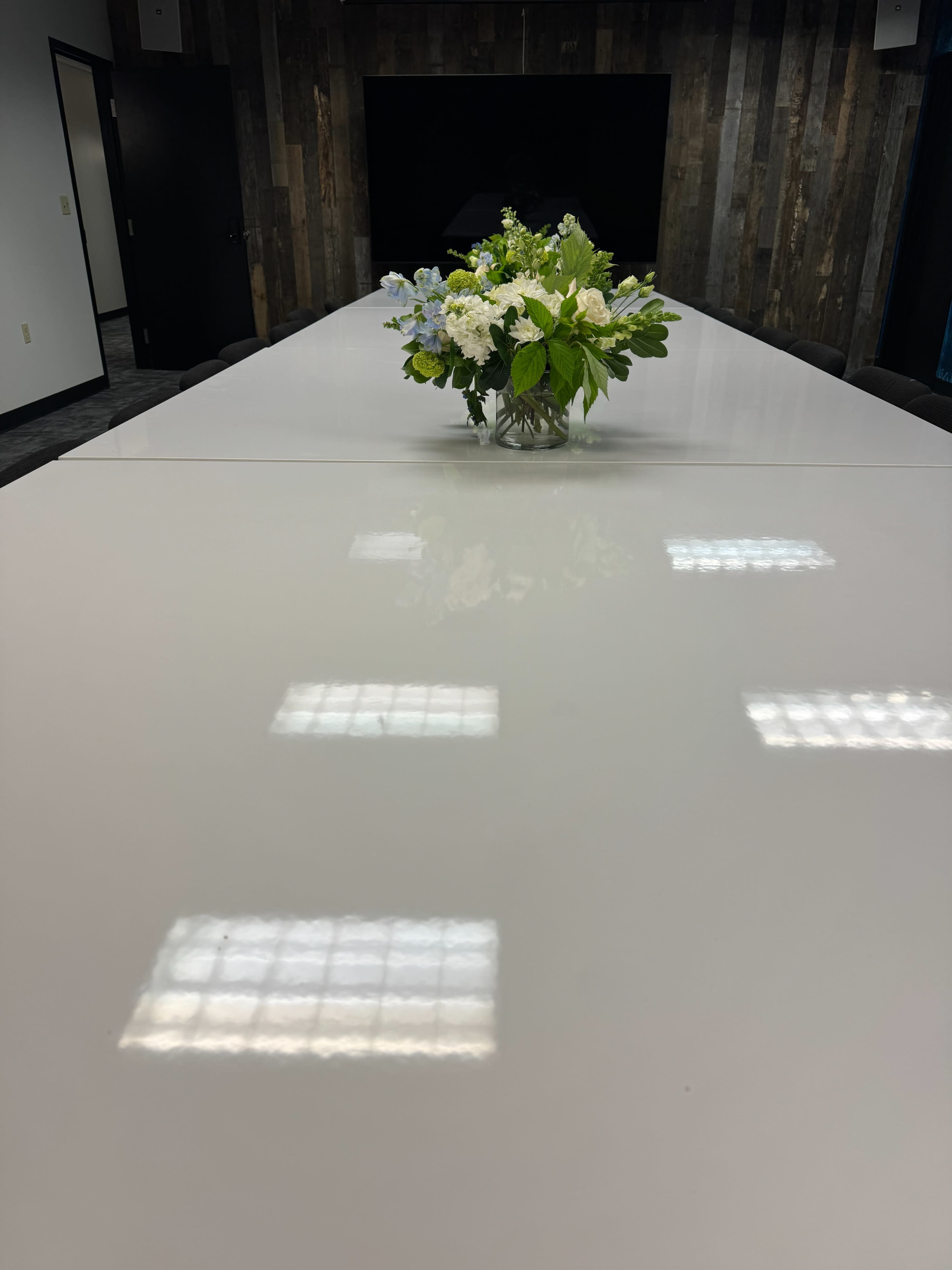 A long, white conference table is set with a bouquet of flowers in the center, reflecting overhead lights and surrounded by dark chairs.