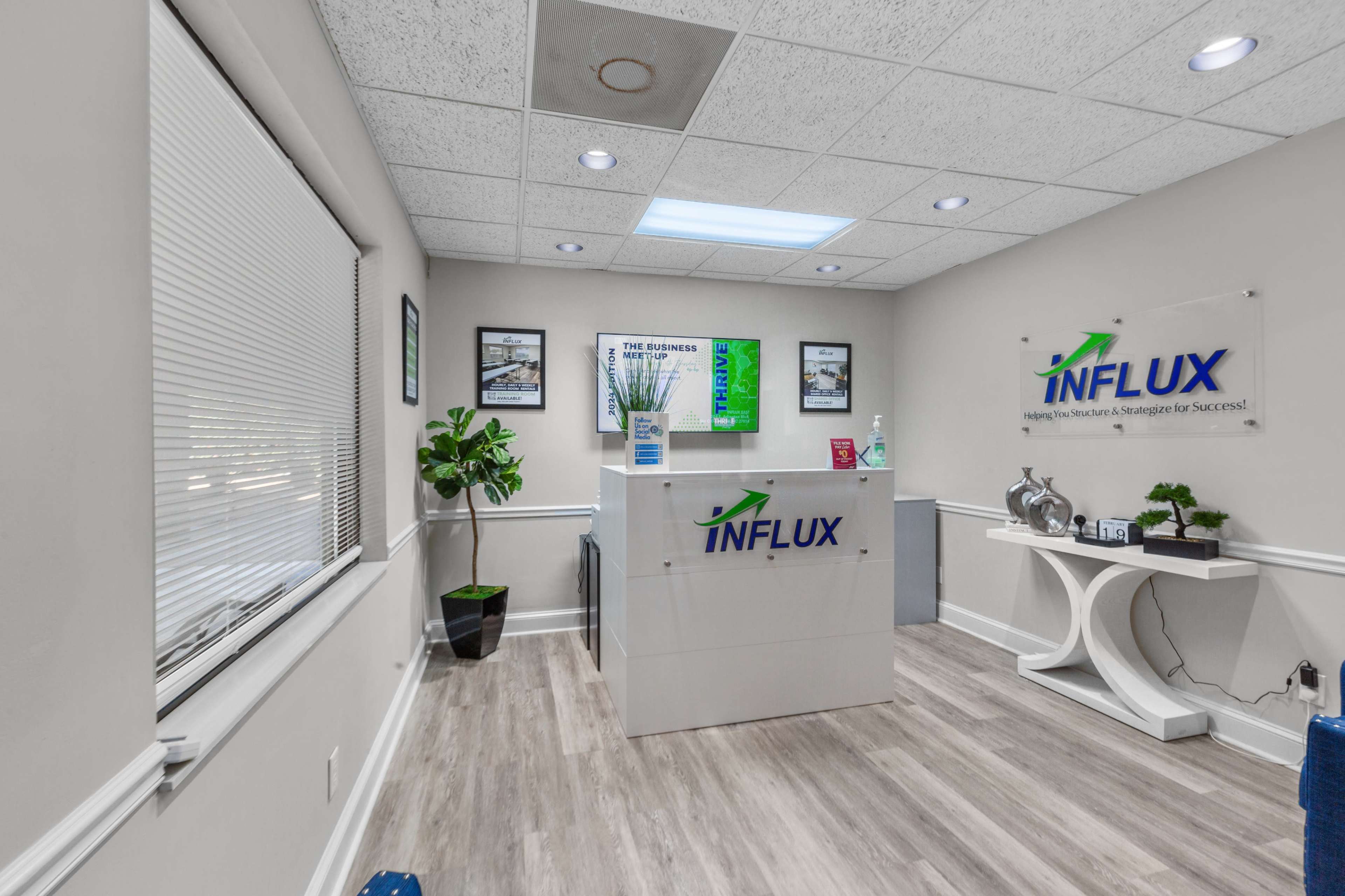 A modern office reception area with a front desk marked "INFLUX," surrounded by framed pictures and a potted plant.