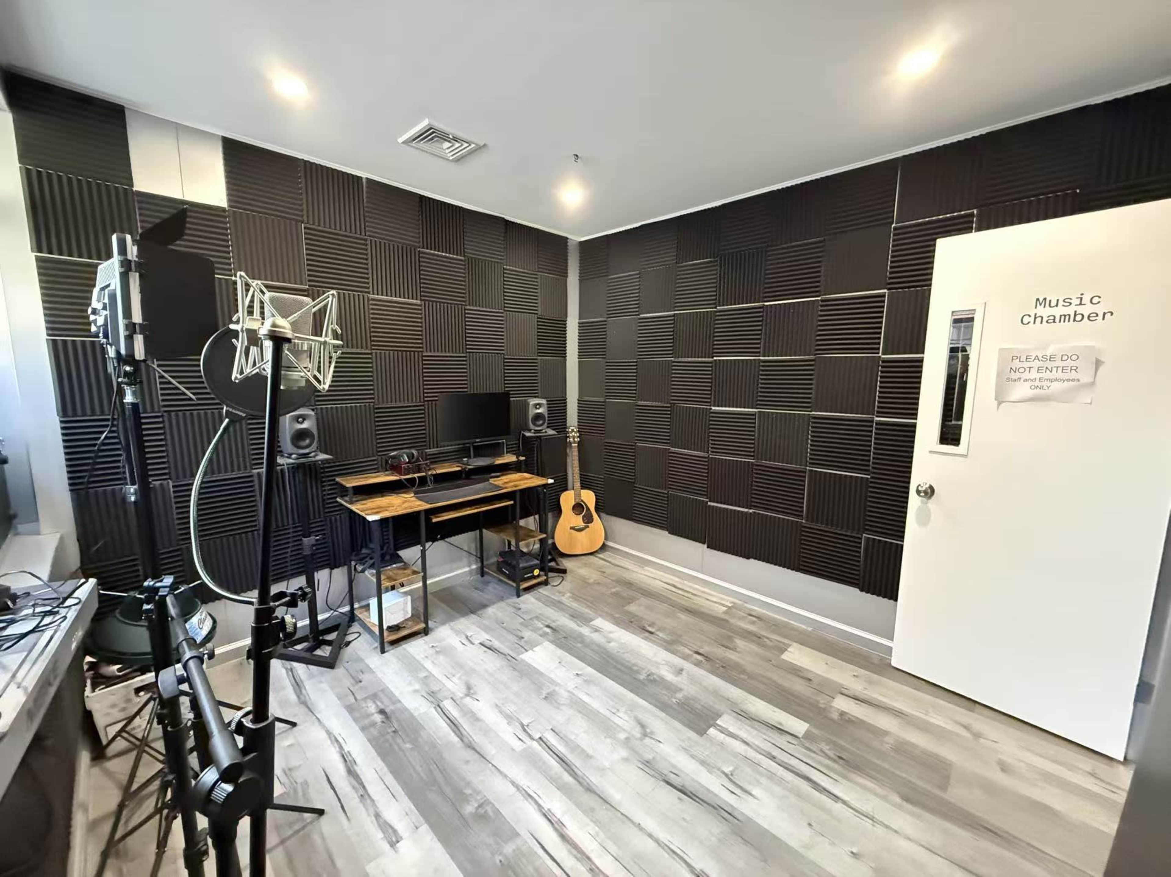 The image shows a small music recording studio with acoustic foam panels on the walls, a desk with a computer and speakers, and an acoustic guitar resting against the wall.