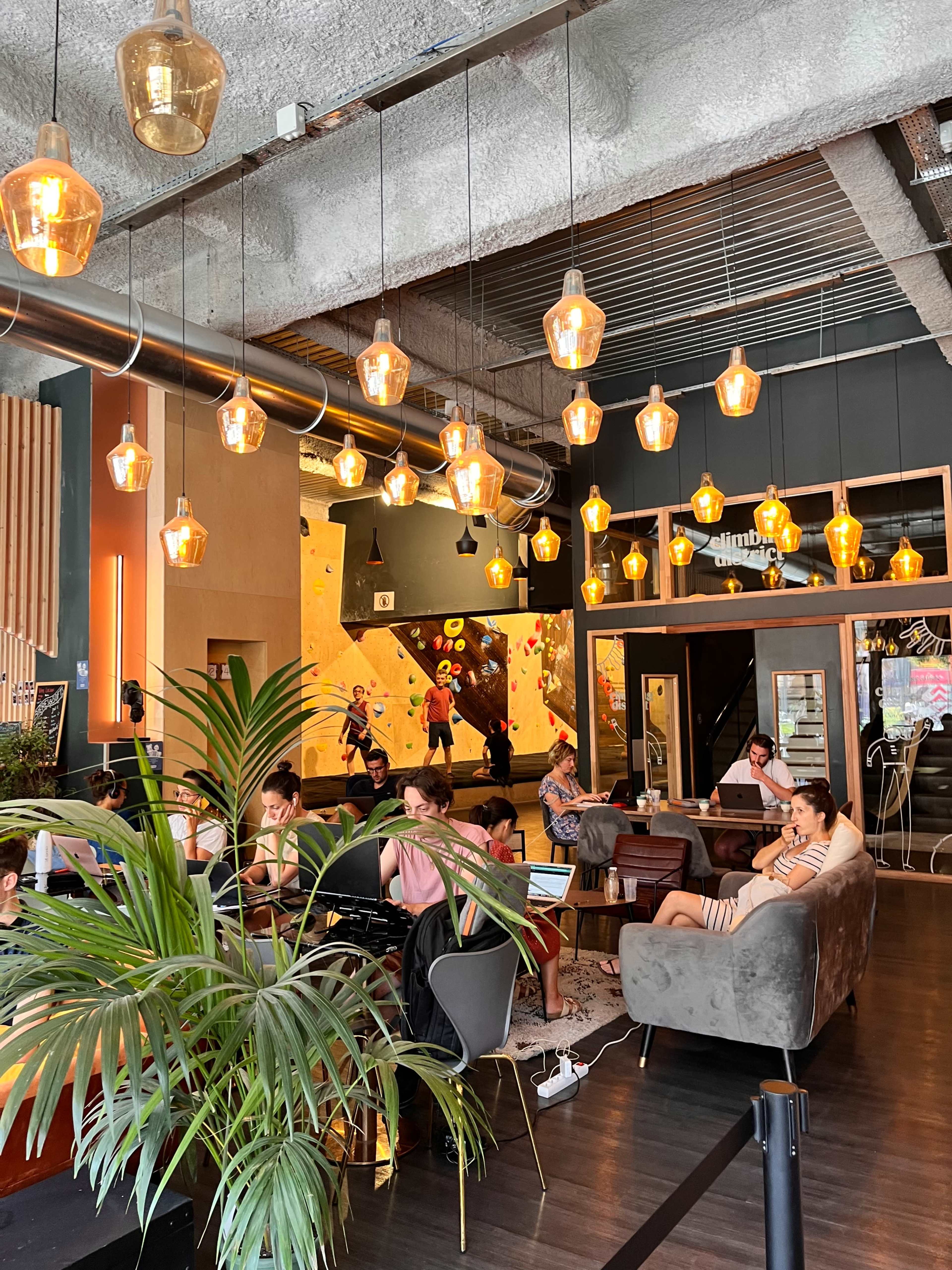 A modern café interior with hanging pendant lights, a mix of seating arrangements, and patrons engaged in various activities.