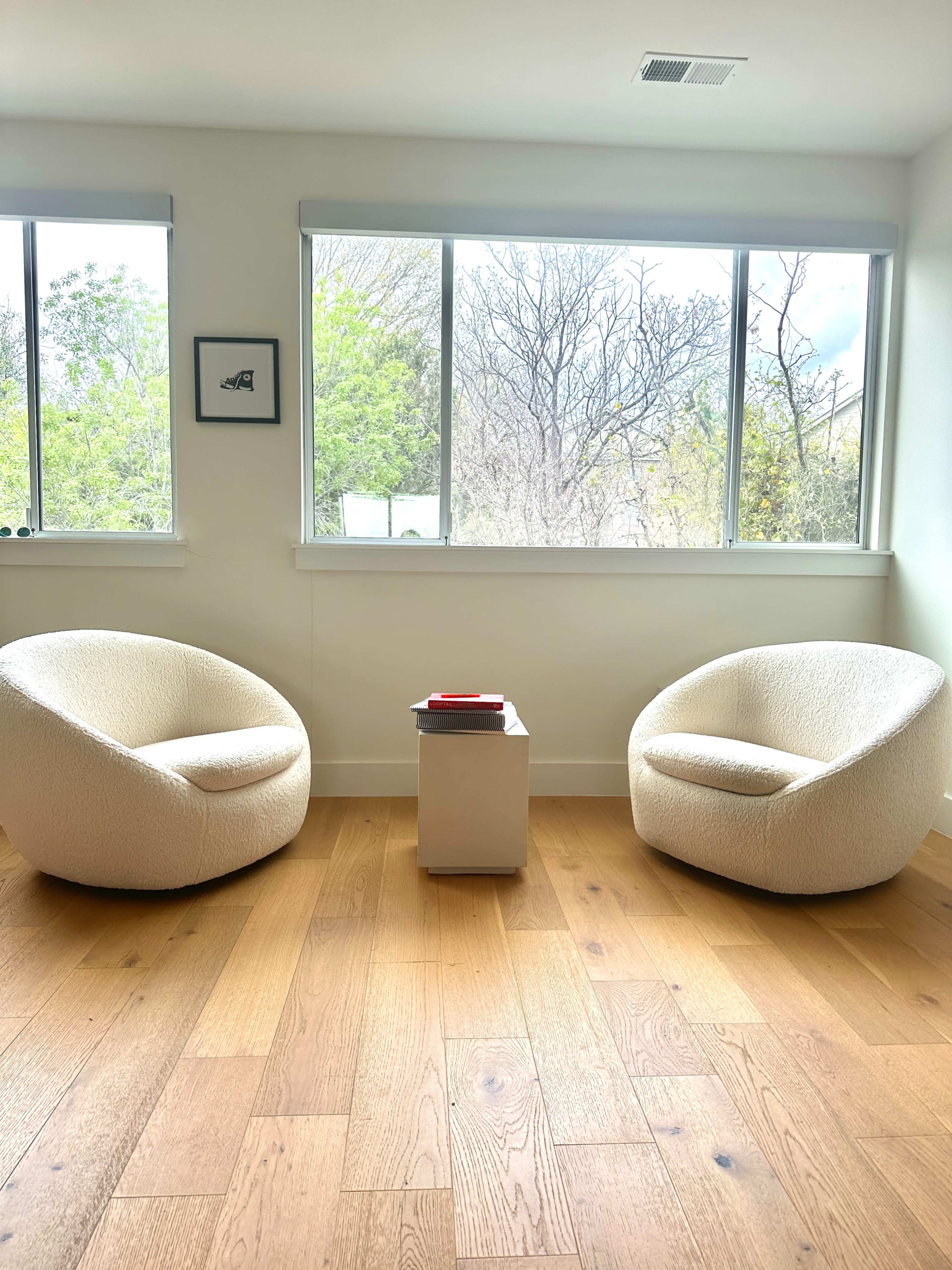 Two rounded chairs are positioned across from each other beside a small square table in a well-lit room with large windows overlooking greenery.