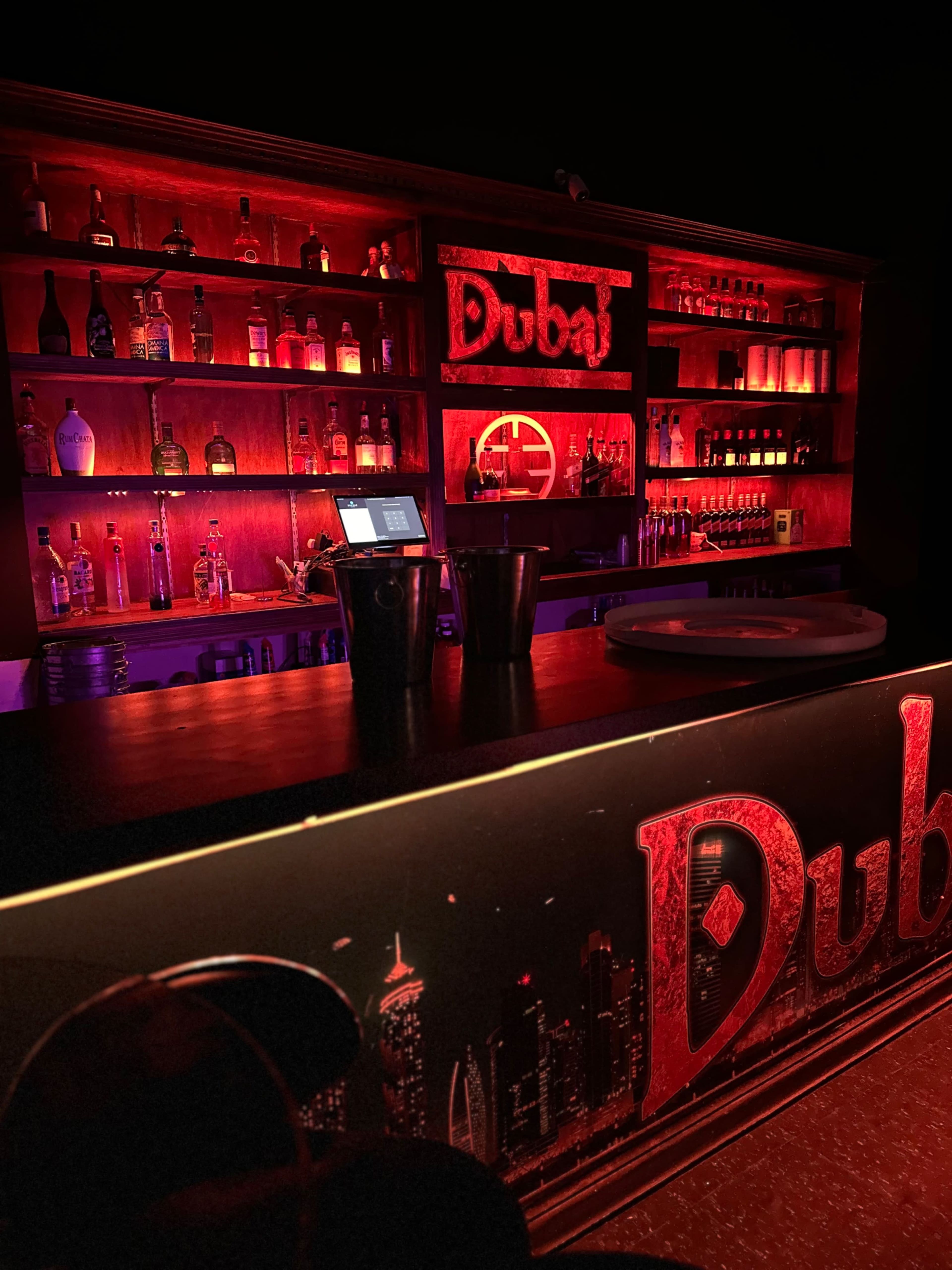 The image shows a dimly lit bar with red lighting, displaying a variety of liquor bottles on shelves behind the counter.