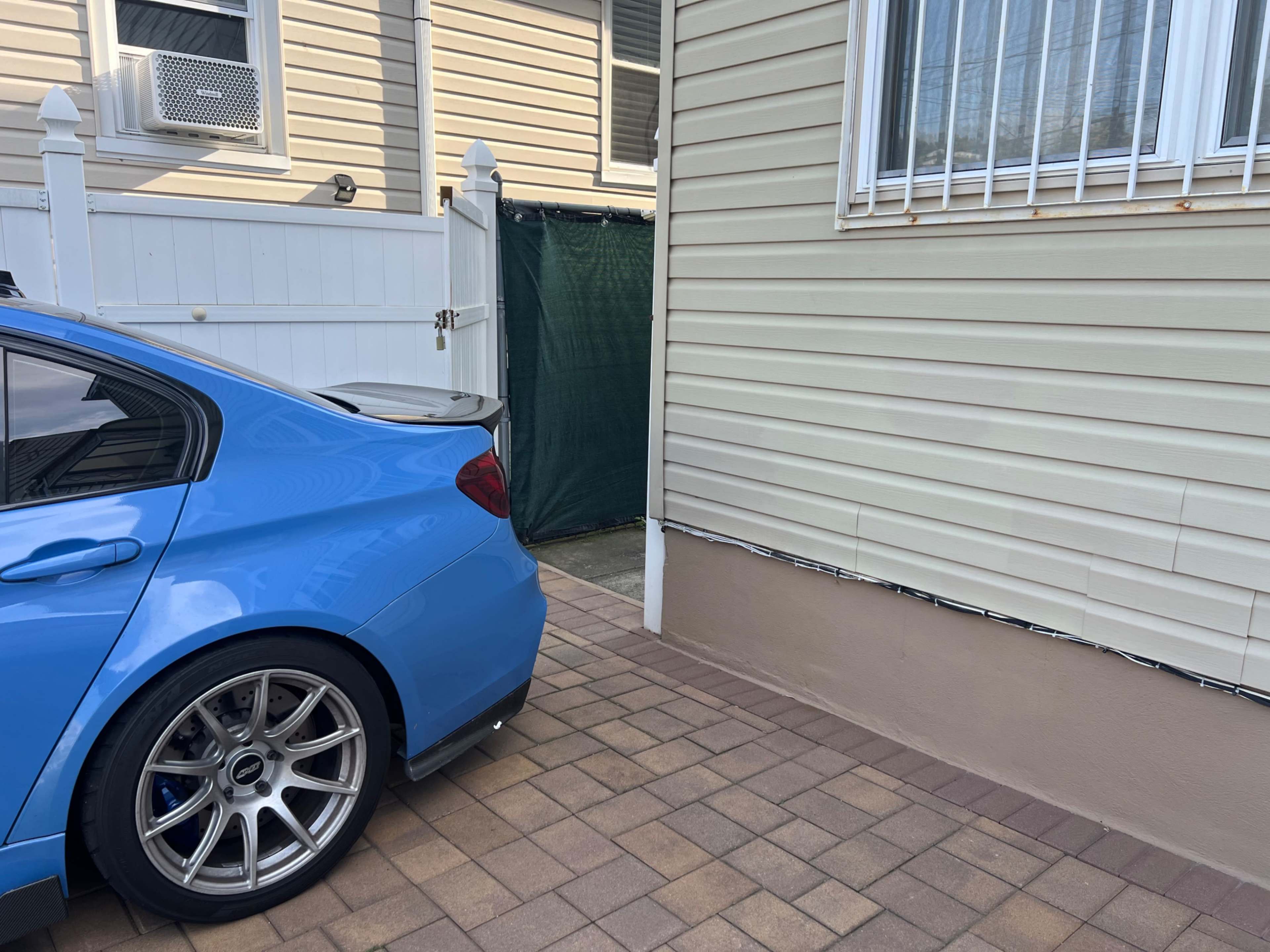 A blue car is parked next to a house, with a green fence and a door visible in the background.