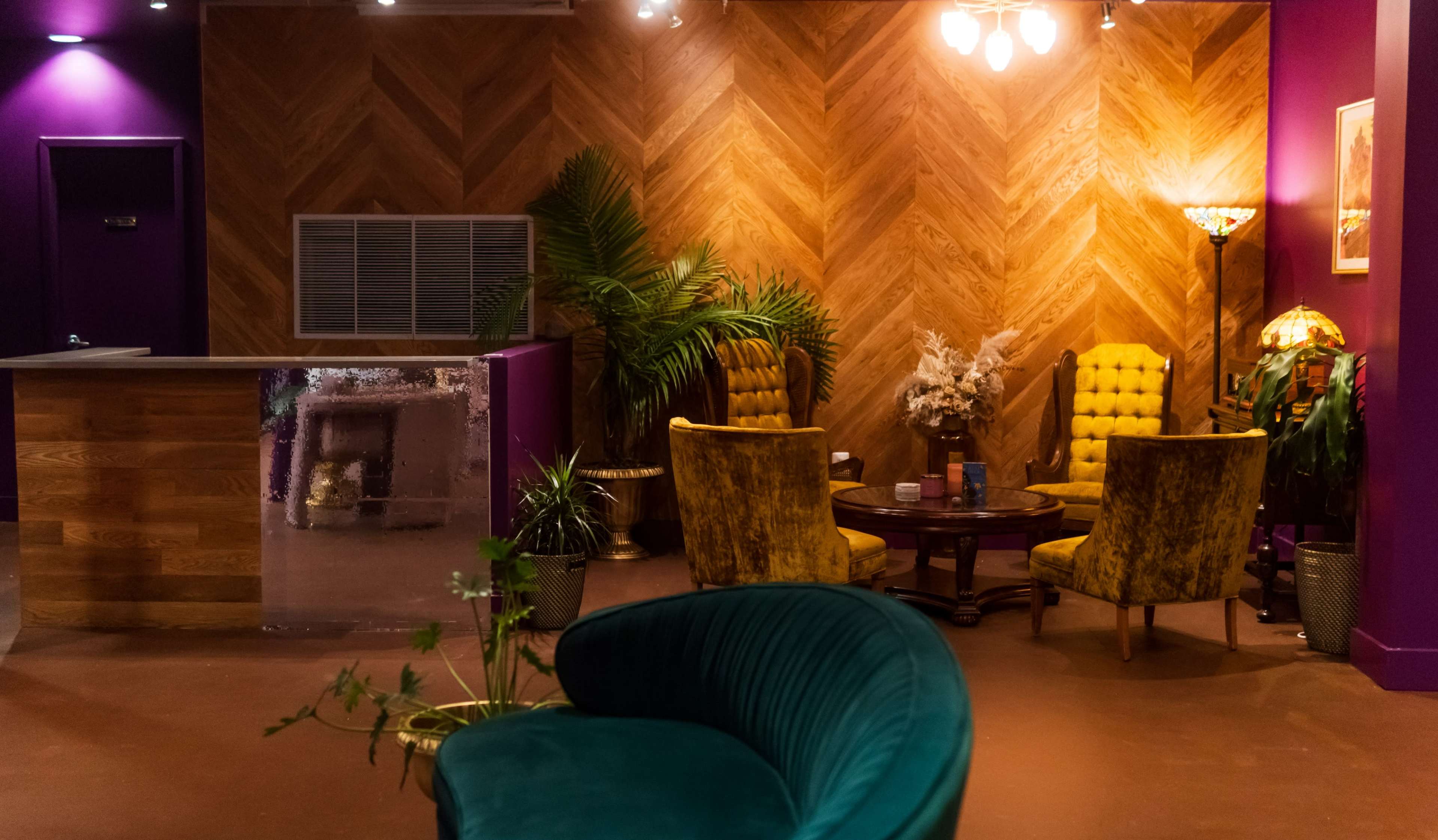 The scene features a modern lounge area with a wooden reception desk, two yellow upholstered chairs, a round table, and decorative plants against a backdrop of herringbone wood paneling and purple walls.