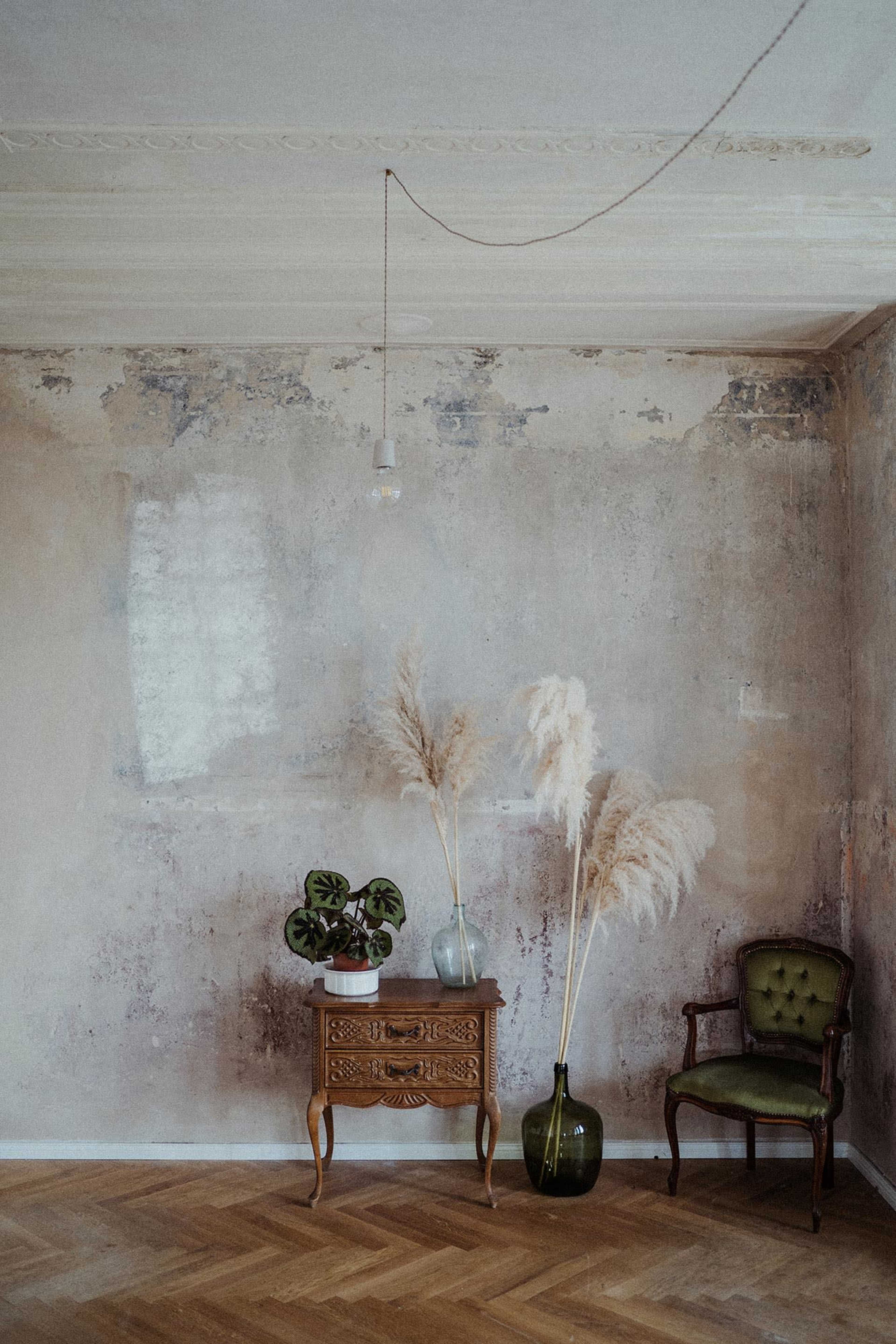 A room with a weathered wall, featuring a wooden table, a green chair, and decorative plants in vases.