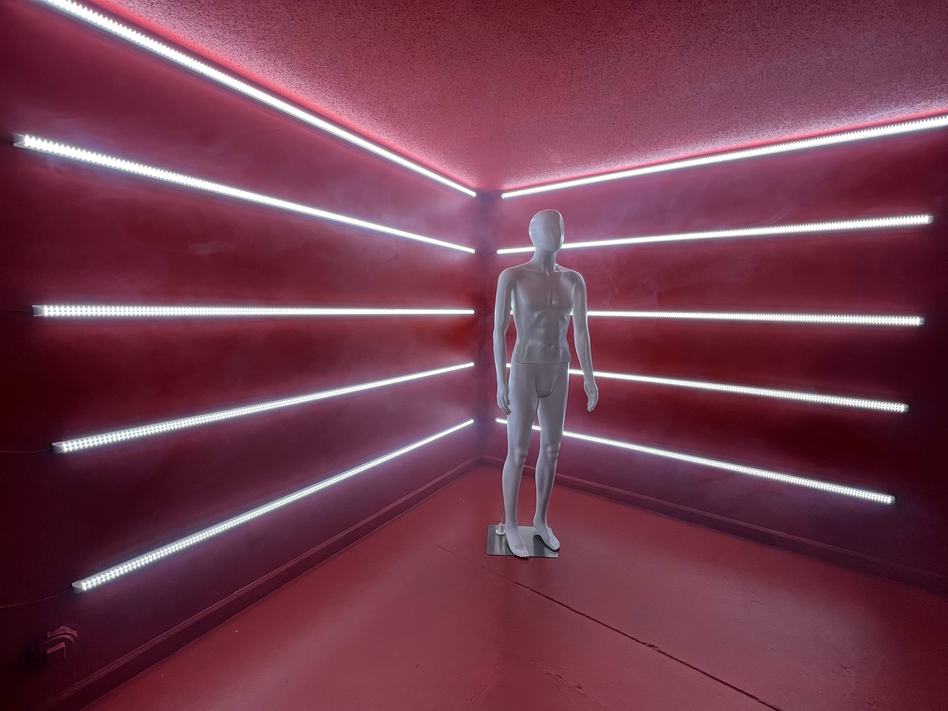 A white mannequin stands in a dimly lit room with red walls and illuminated by horizontal strips of white LED lights.