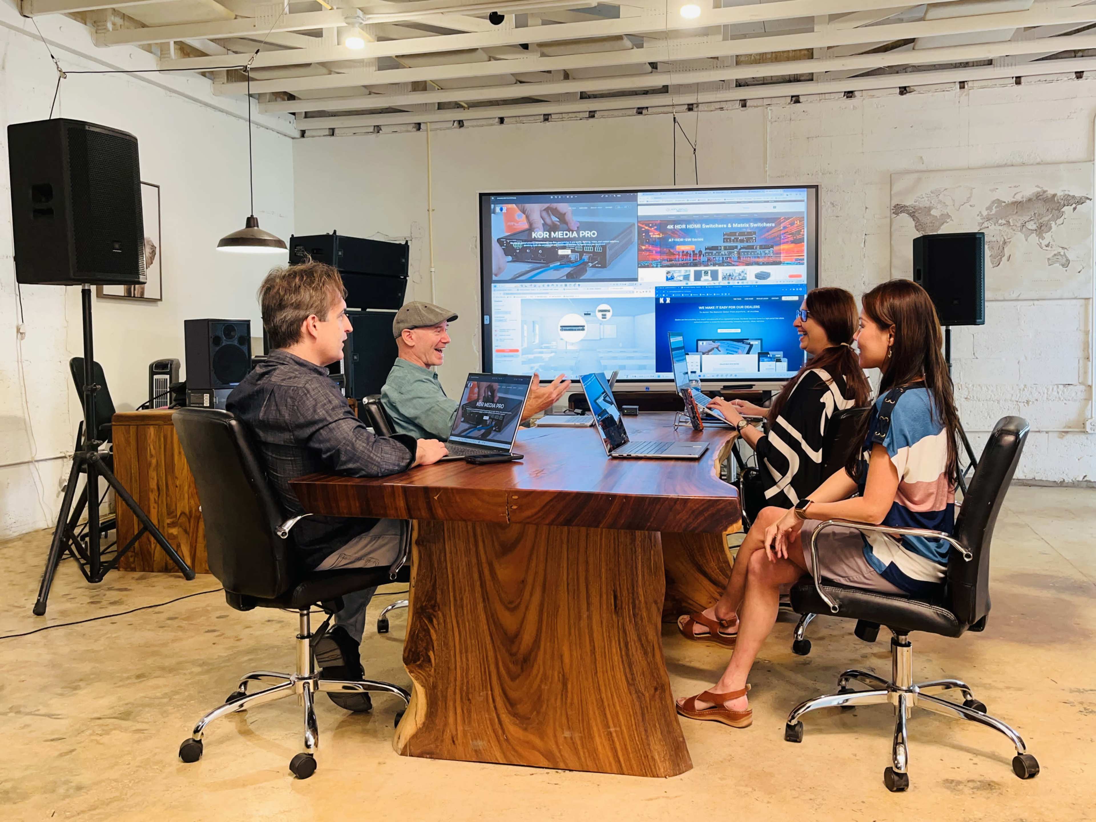 A group of four people is seated around a wooden conference table, engaging in a discussion while using laptops in a modern office space.