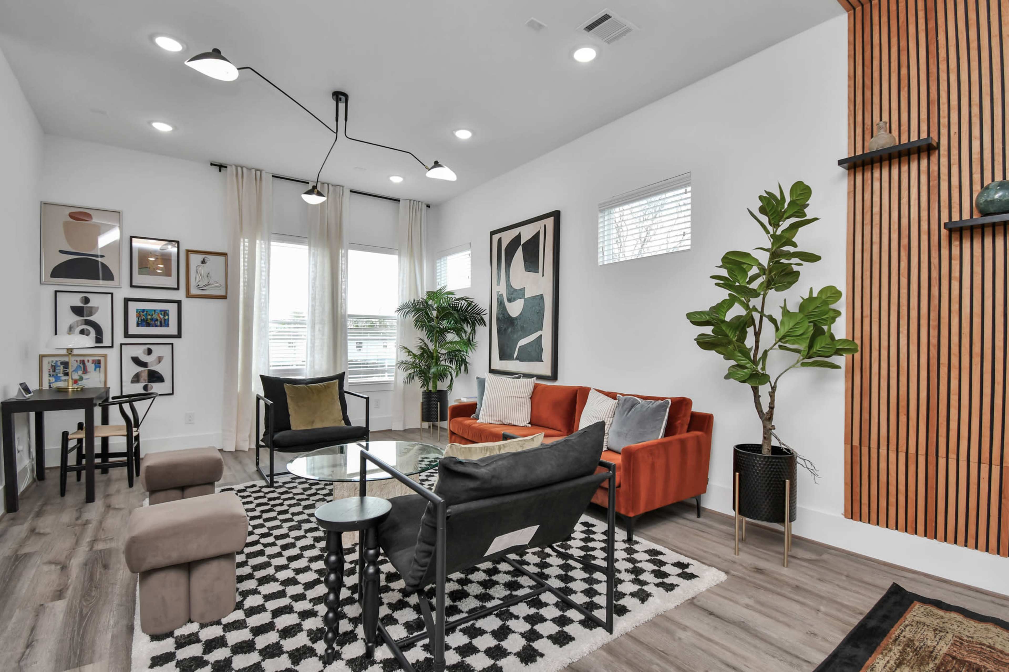 The living room features a mix of modern furniture, including an orange sofa, a black chair, and a patterned rug, with large windows that allow natural light to fill the space.