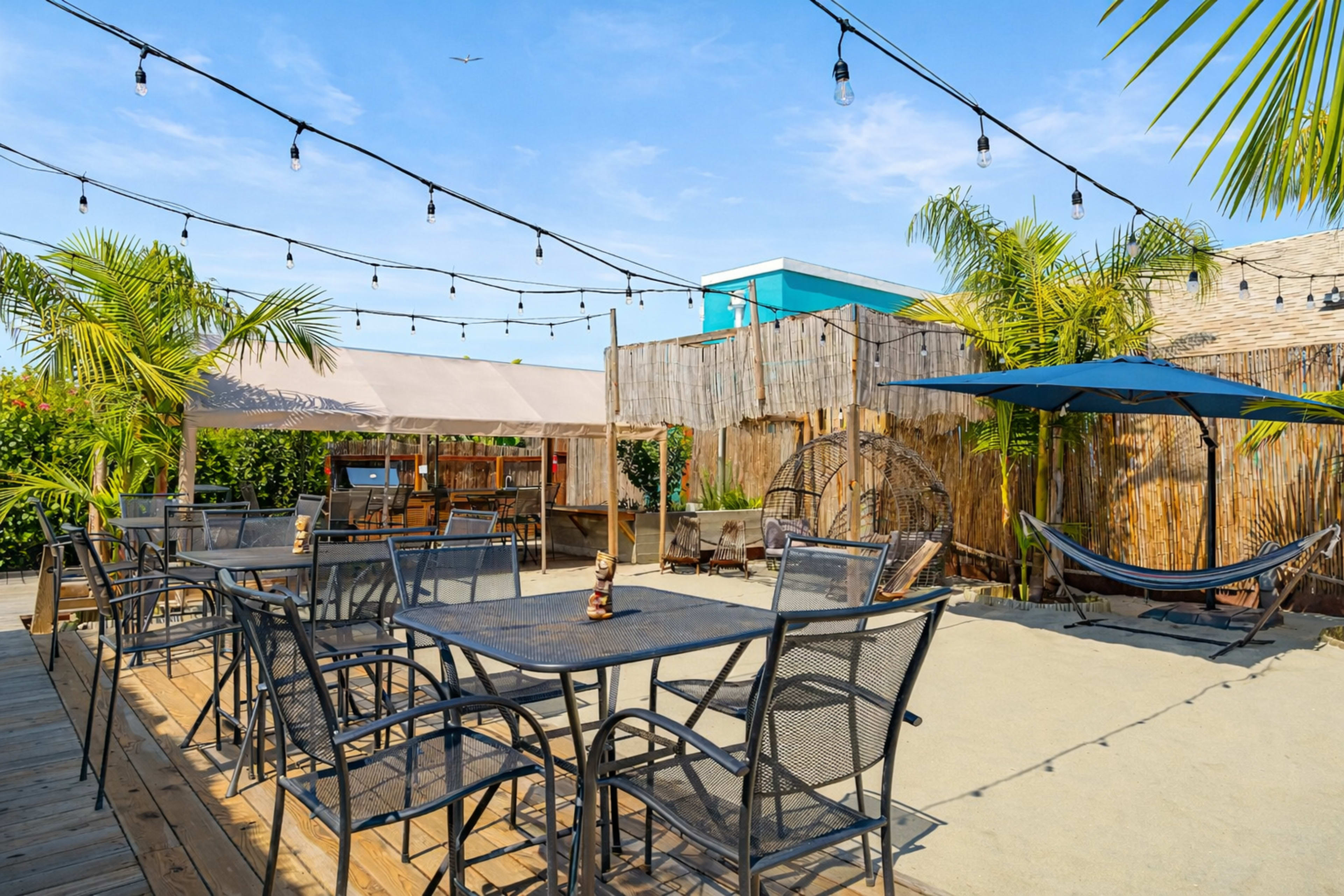 The image shows a vibrant outdoor seating area with black metal tables and chairs, surrounded by palm trees and string lights, on a sandy surface.