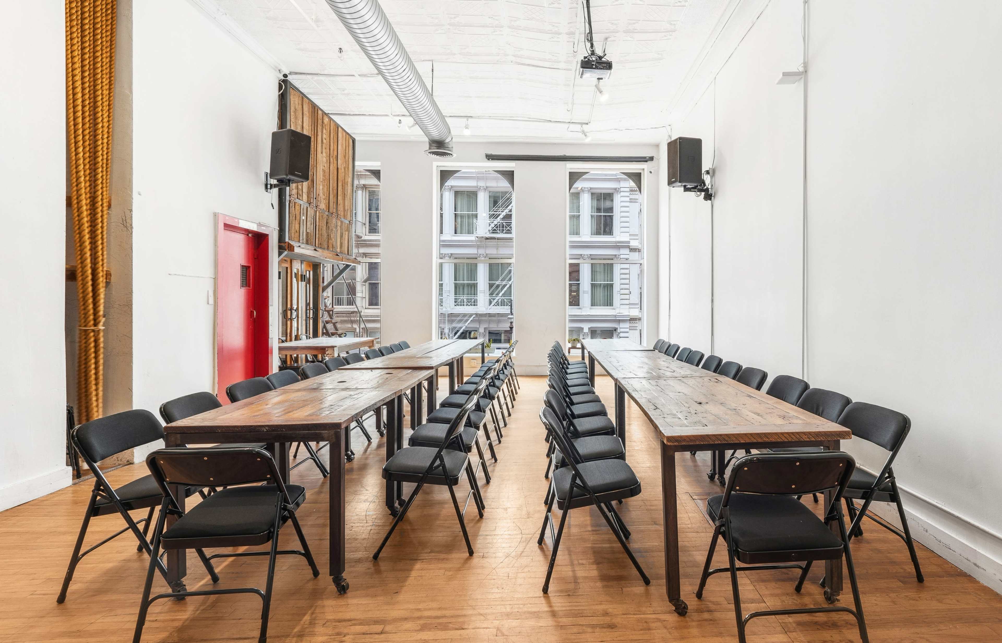 A spacious room features two long wooden tables arranged in parallel with black folding chairs positioned around them, illuminated by natural light from large windows.