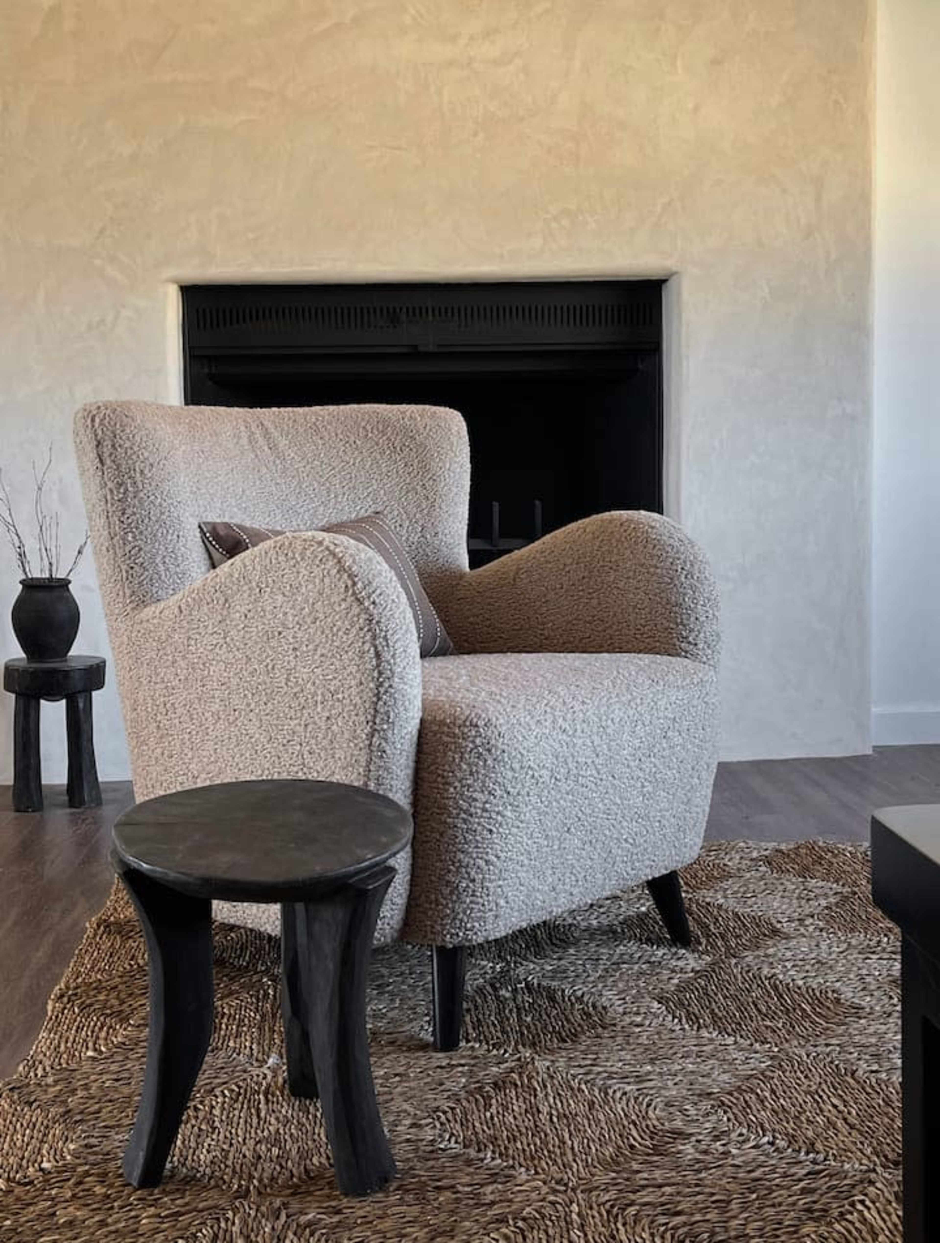 A textured beige armchair sits beside a small black pedestal table on a woven rug in a minimalist living room with a blank wall and a fireplace.