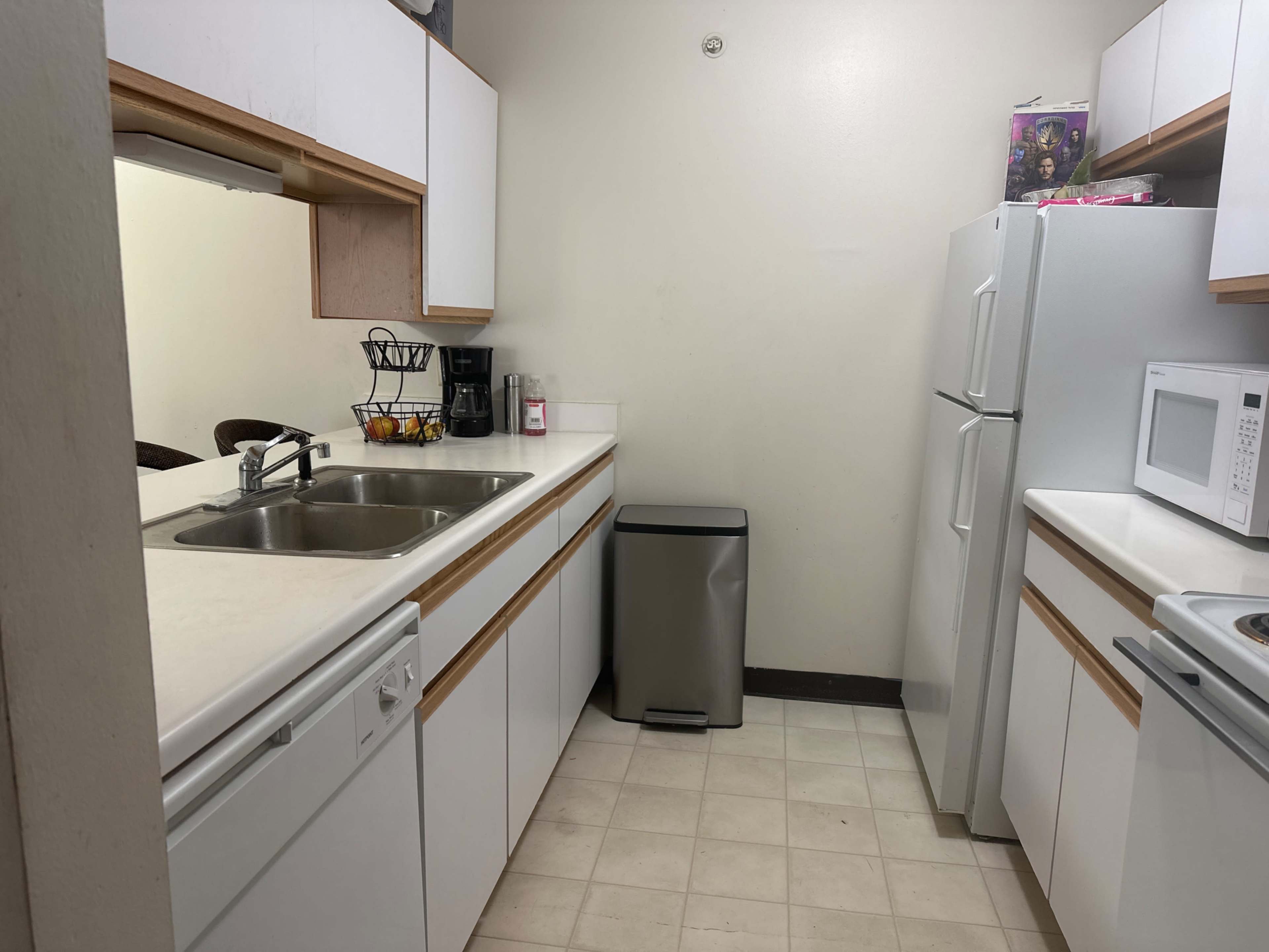 A small kitchen features a double-basin sink, a refrigerator, a microwave, and a stove, with a trash can and a coffee maker on the counter.