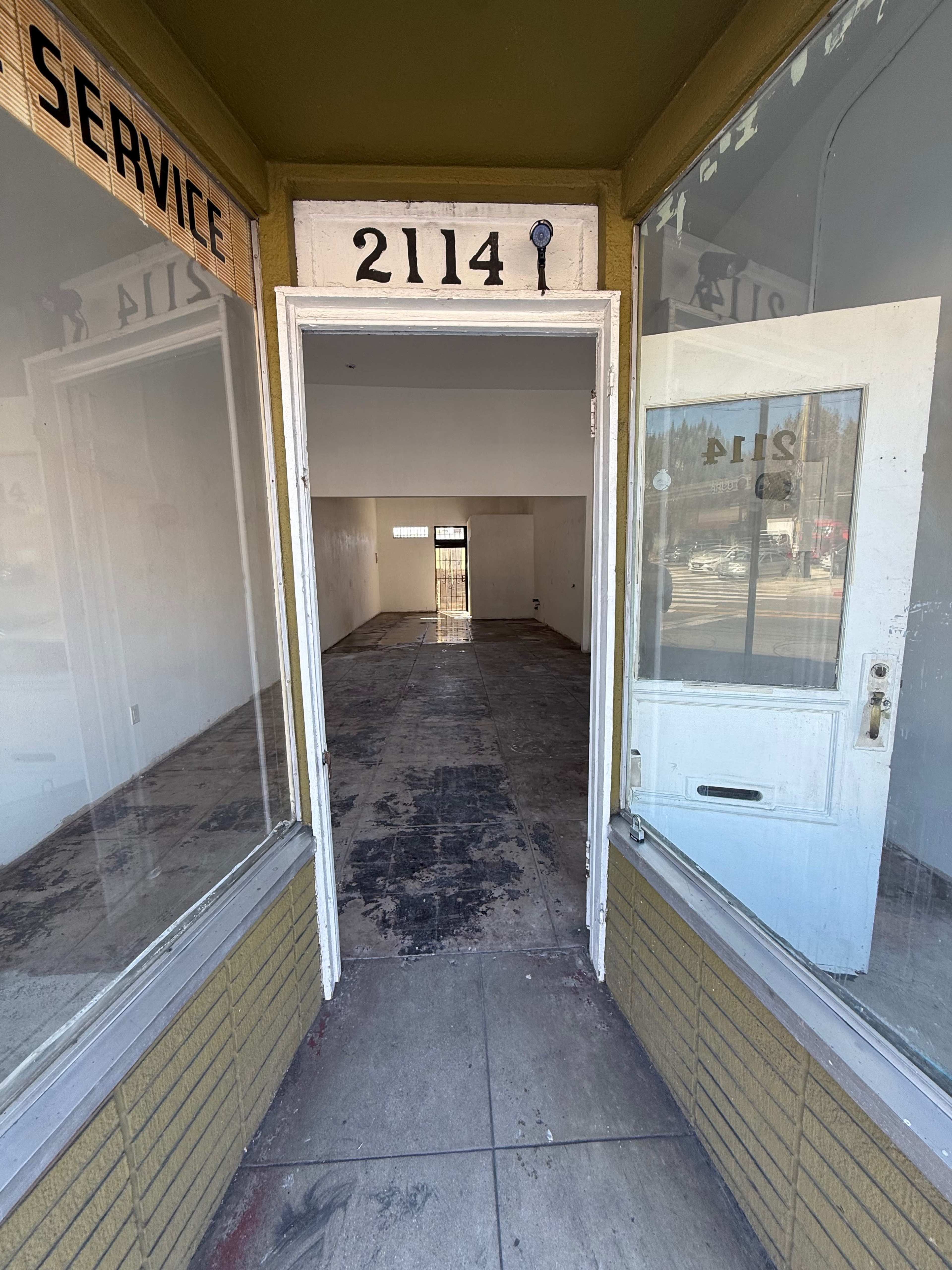 The image shows the entrance to a vacant commercial space with the number "2114" displayed above the door and empty interior walls and floor visible through the front windows.