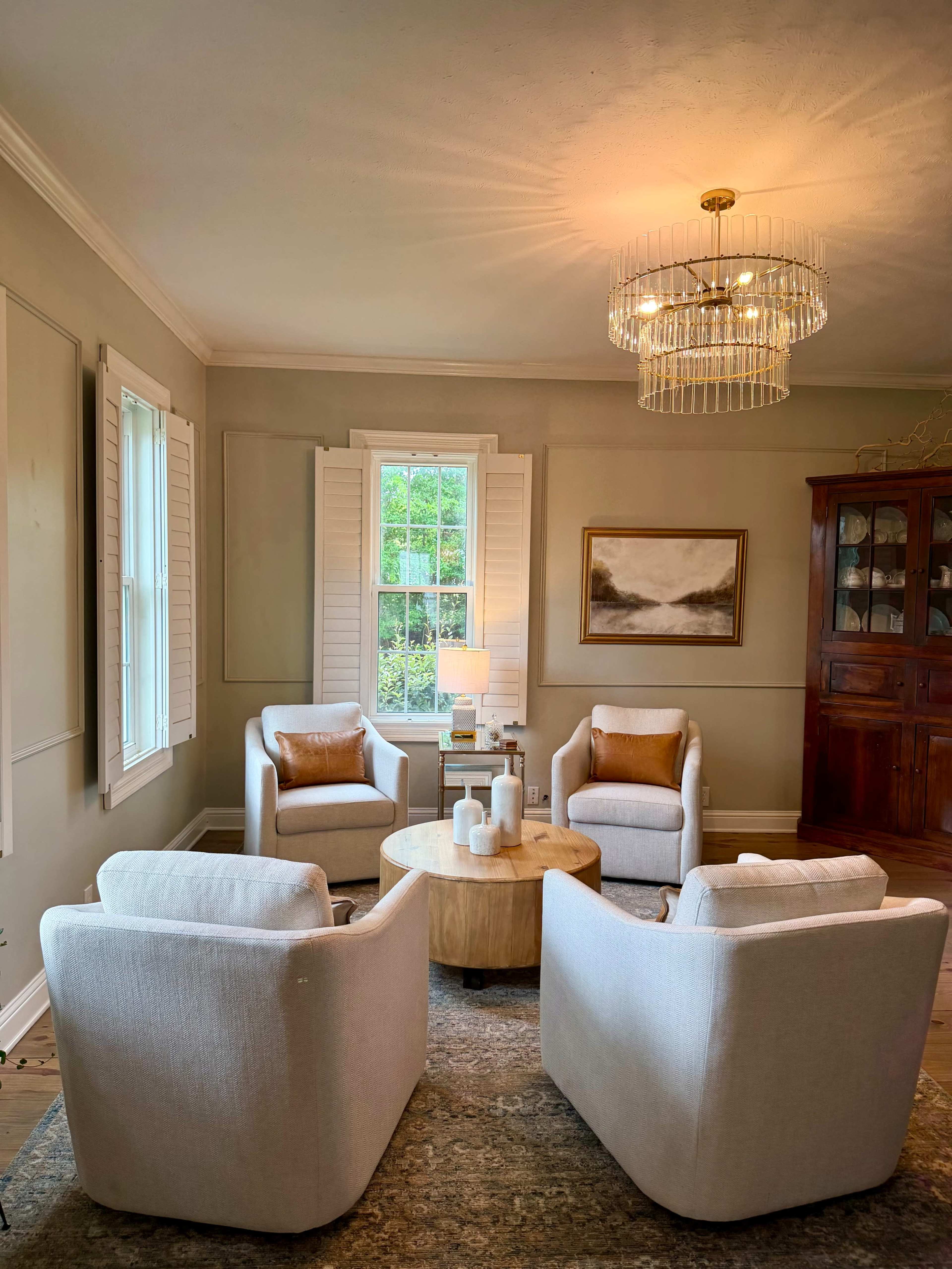 The image shows a cozy living room with four upholstered armchairs arranged around a circular wooden coffee table, illuminated by a chandelier.