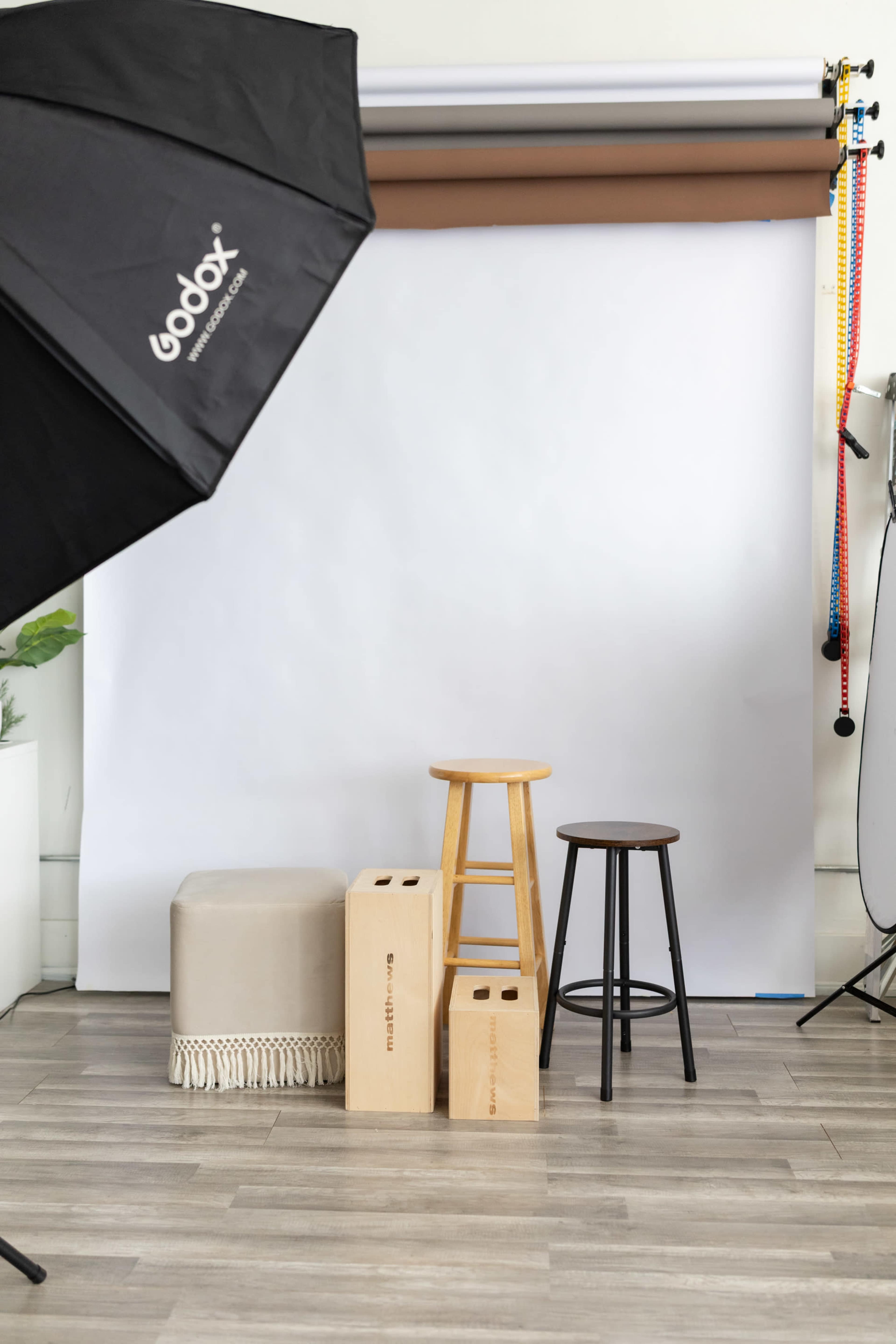 The image shows a minimalistic photo studio setup with a white backdrop, three stools of different heights, and a softbox lighting umbrella.