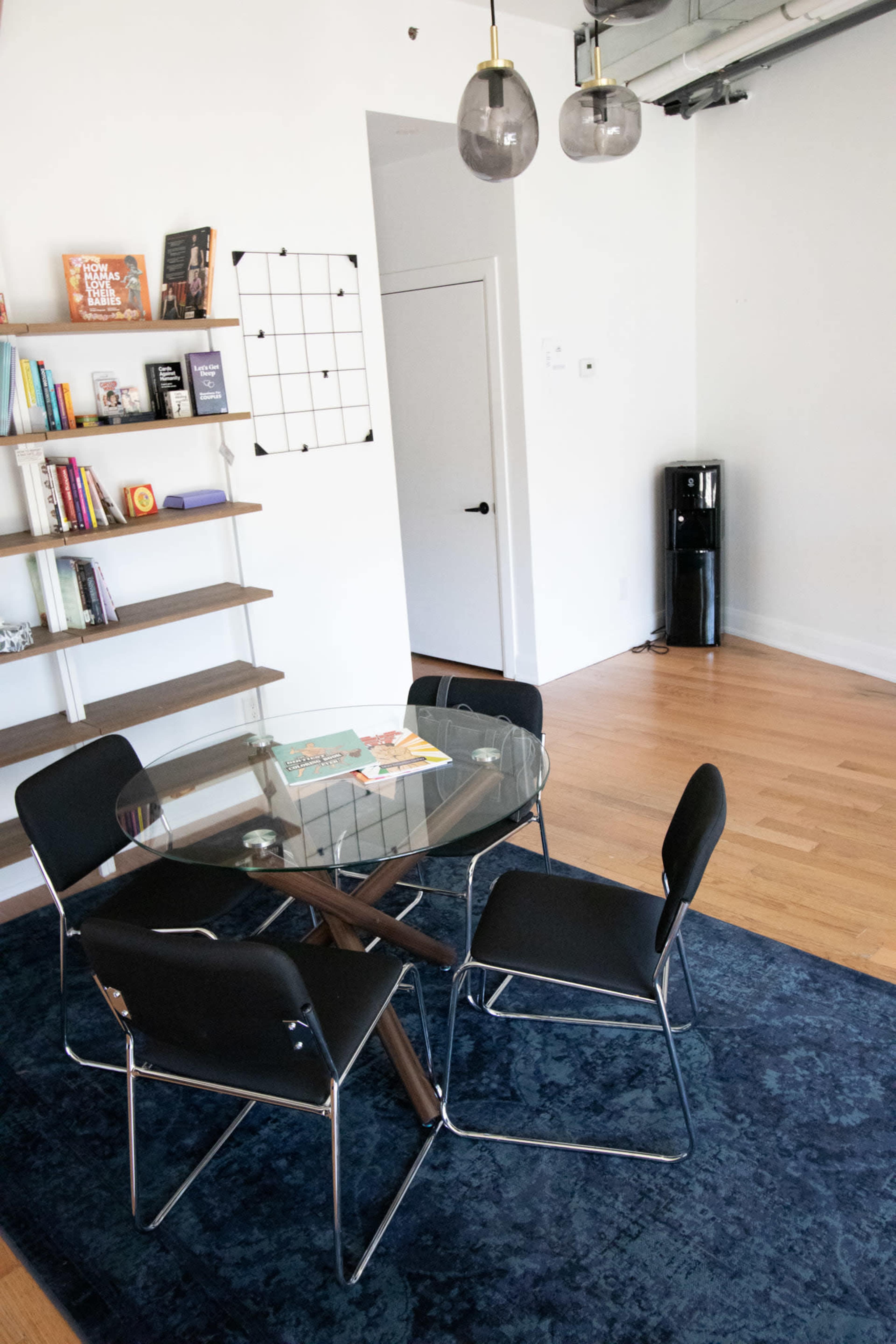 A glass dining table surrounded by four black chairs is set in a brightly lit room with bookshelves and a black refrigerator in the corner.